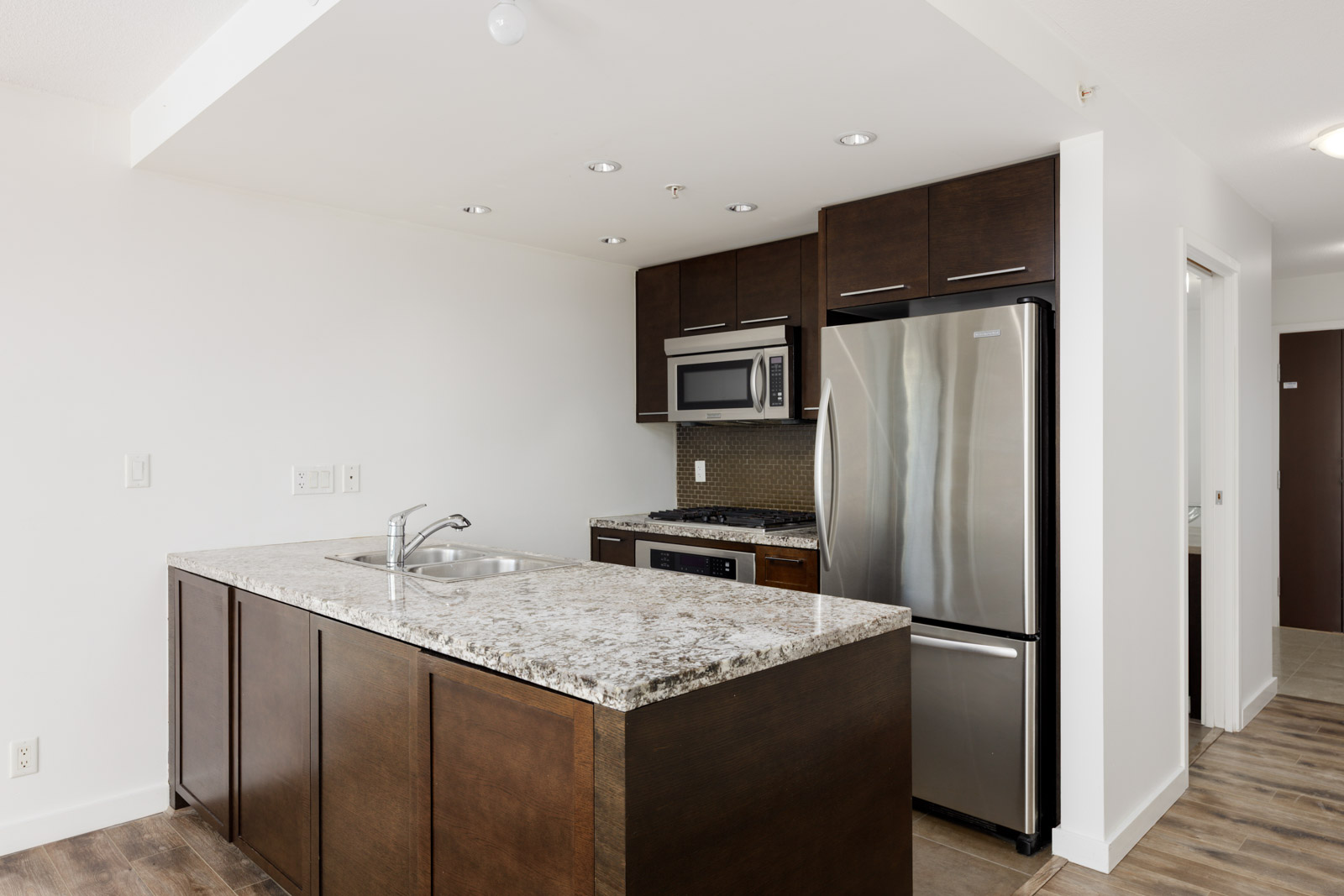 Modern kitchen with dark wood cabinets, stainless steel appliances, granite countertop island with a sink, and light-colored walls and flooring.