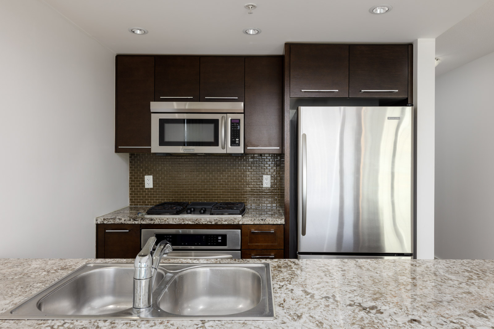 Modern kitchen with dark wood cabinets, stainless steel refrigerator, microwave, stove, oven, and a granite countertop with a double sink in the foreground.