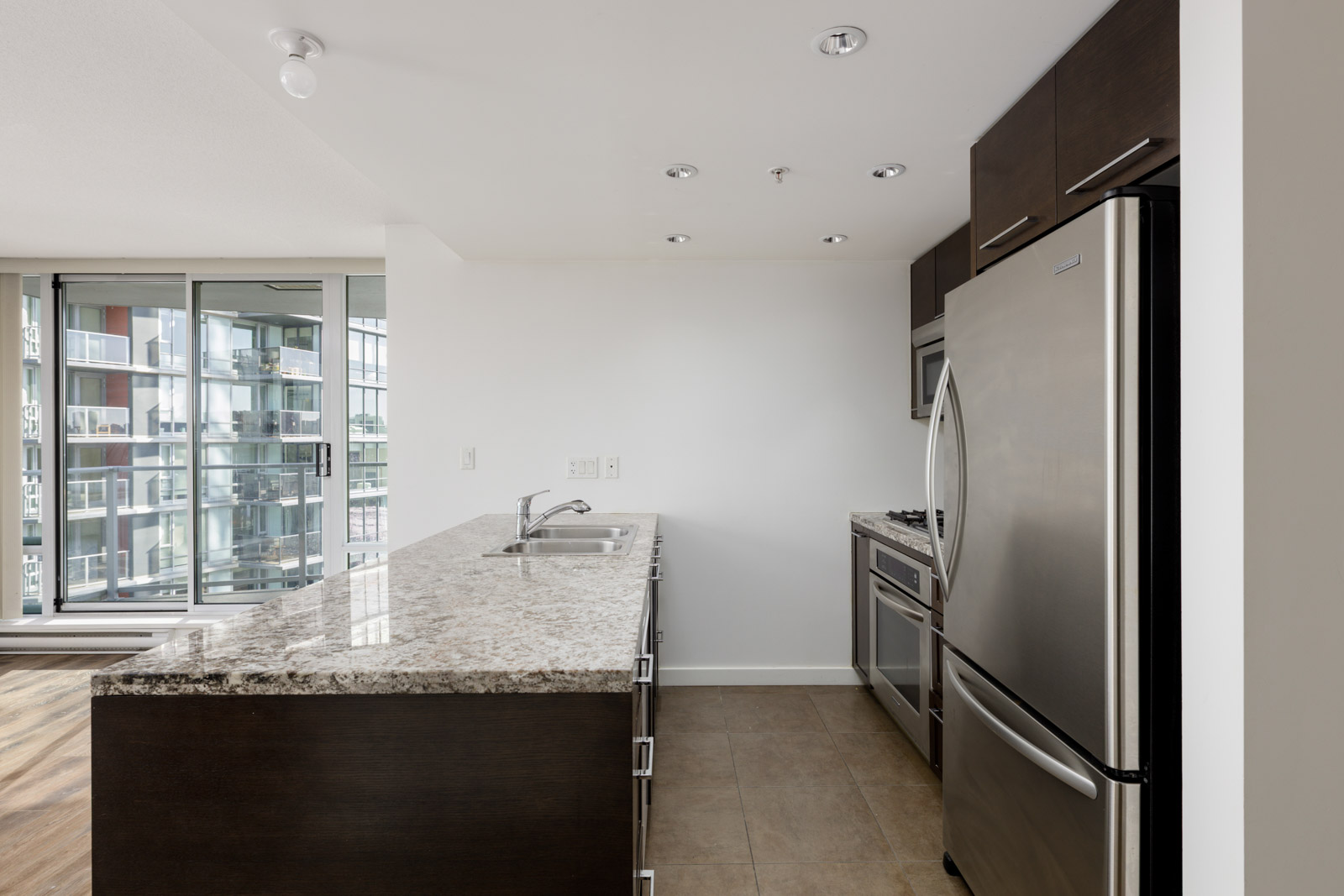 Modern kitchen with stainless steel appliances, granite countertop island with sink, dark cabinets, and large windows overlooking a balcony and cityscape.