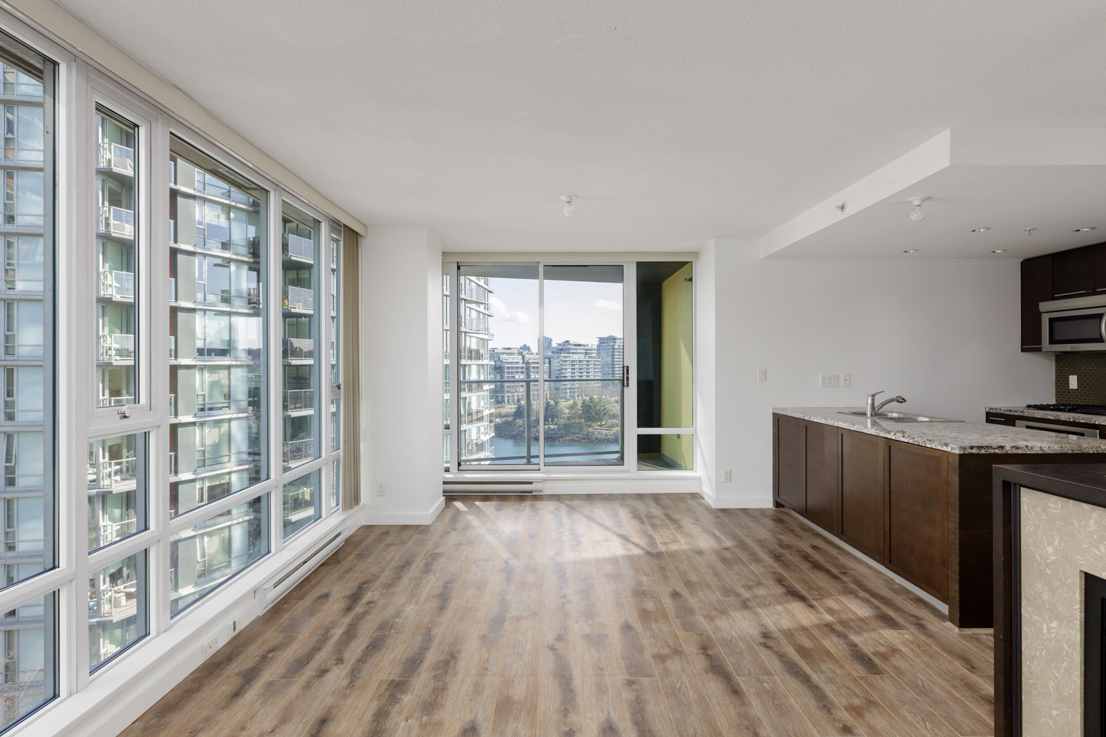 A modern, unfurnished apartment with large windows, wood flooring, an open kitchen, and a balcony overlooking city buildings and water.