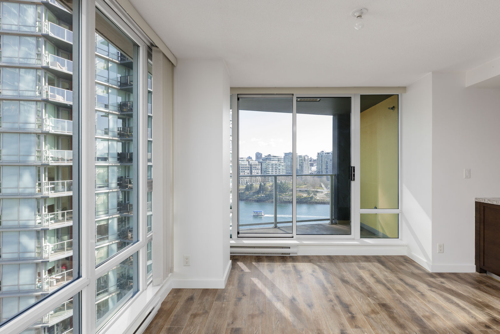 Bright modern apartment with large windows, wood flooring, and a balcony overlooking a body of water and city buildings.
