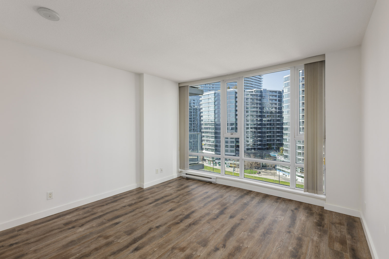 Unfurnished apartment room with white walls, large floor-to-ceiling windows, and wood-style flooring, overlooking a view of modern city buildings.