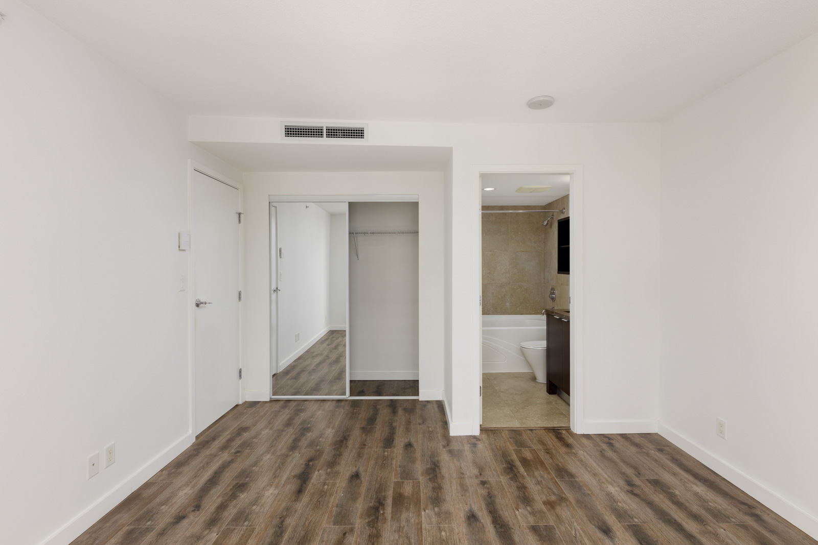Empty room with white walls, wood-style flooring, mirrored closet doors, and an open doorway leading to a bathroom with beige tile.