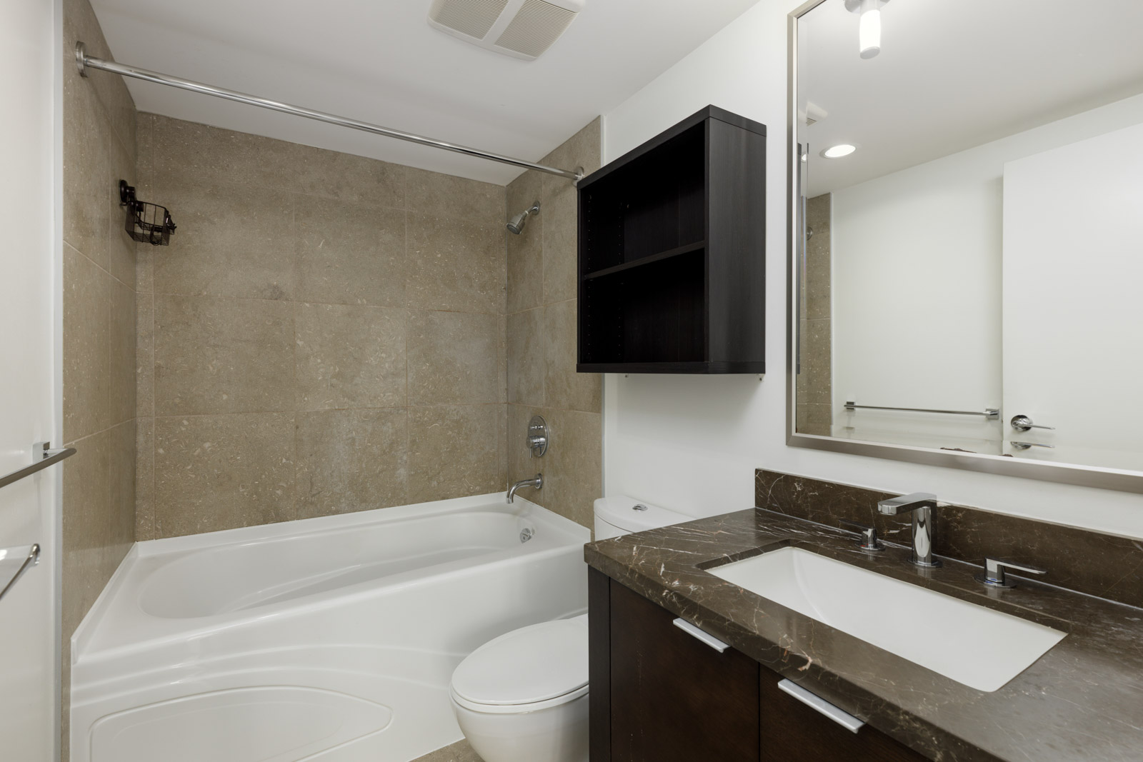 Modern bathroom with a bathtub-shower combo, wall-mounted shelf, toilet, and a sink with a dark countertop and large mirror.