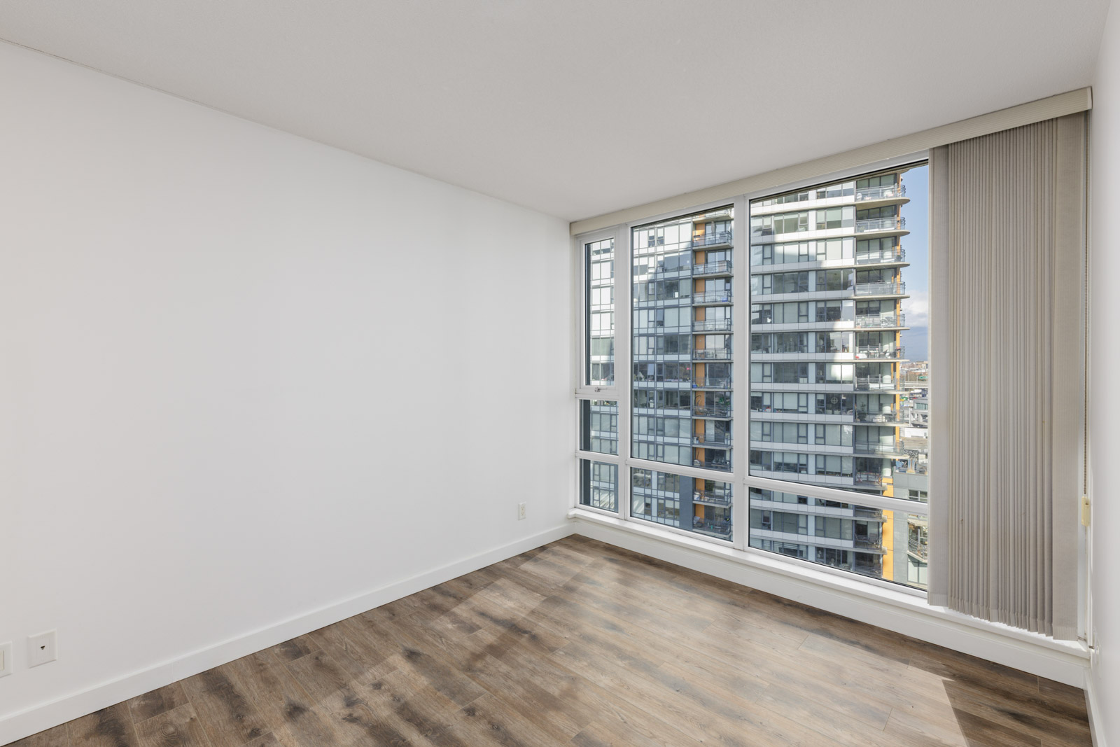 Empty room with white walls, wood flooring, and large floor-to-ceiling windows showing a view of a modern high-rise building outside.