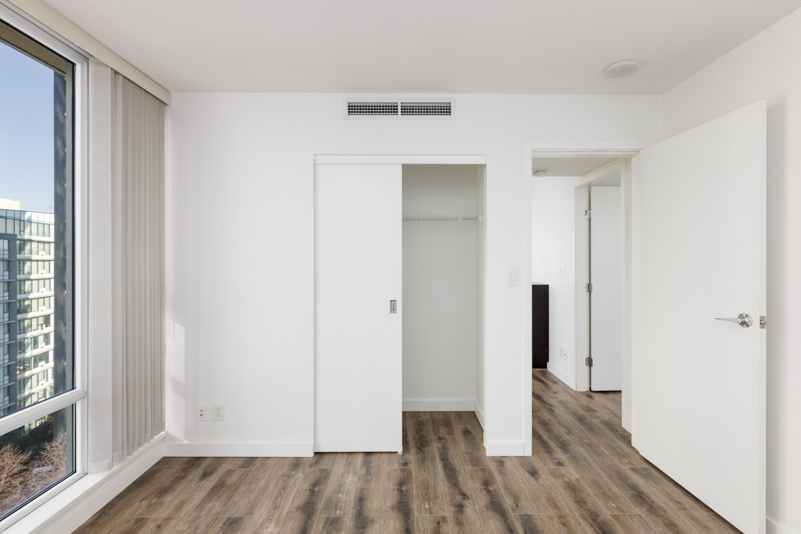 Empty bedroom with wood flooring, sliding closet doors, open entry door, large window with vertical blinds, and white walls.