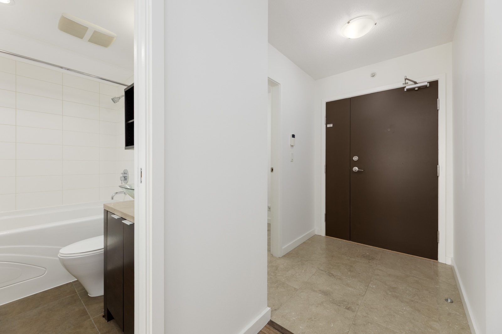View of a bathroom with a bathtub and toilet to the left and a brown apartment entrance door with tile flooring to the right.