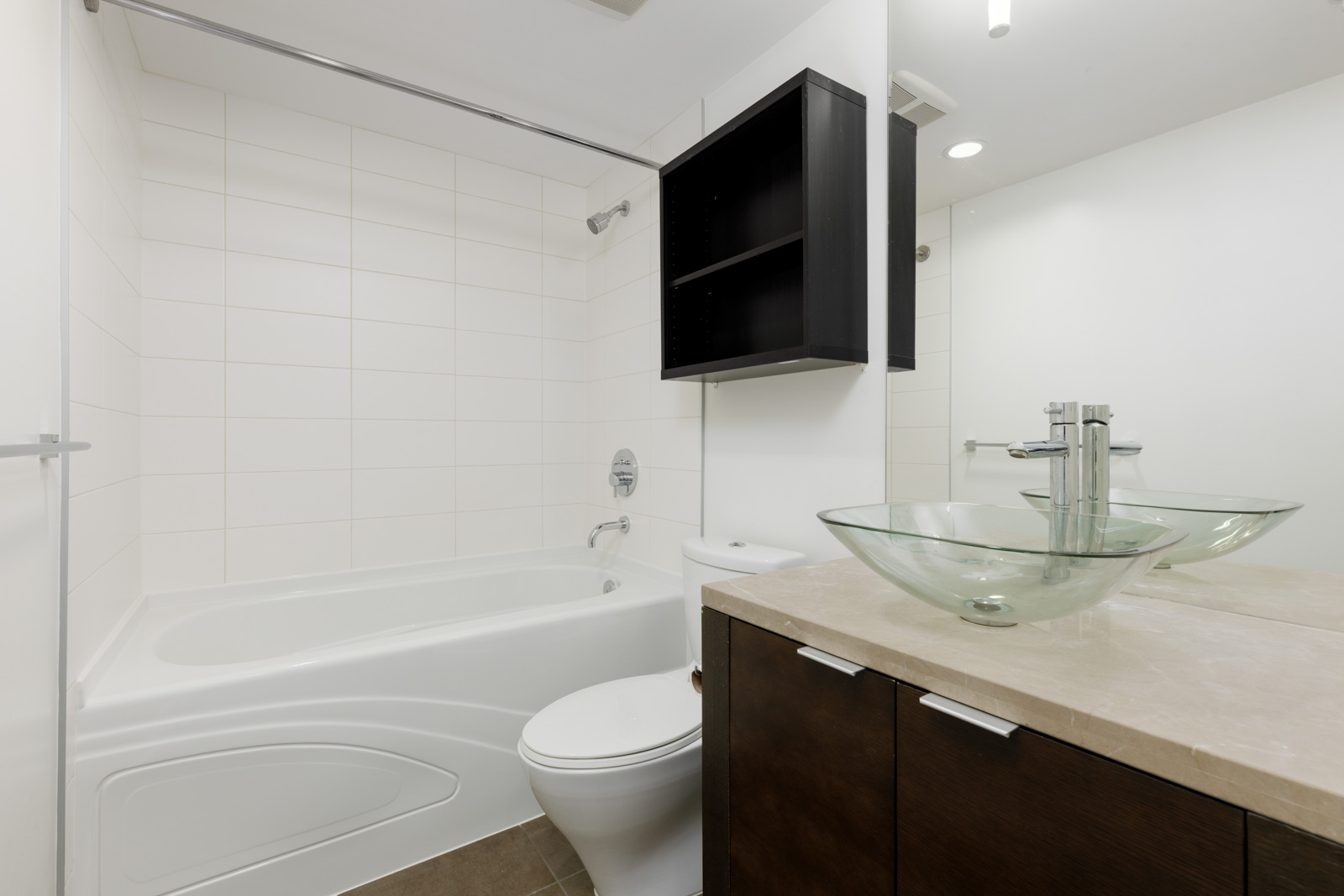 Modern bathroom with a white bathtub, toilet, glass vessel sink on a beige countertop, dark wood vanity, wall-mounted shelf, and white tiled walls.