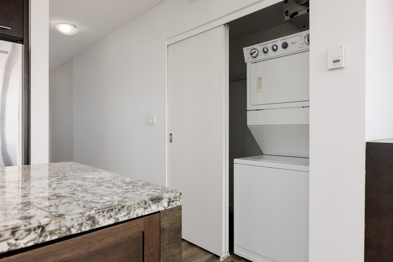 A stacked washer and dryer unit is partially visible behind a sliding closet door in a modern laundry area next to a kitchen with granite countertops.
