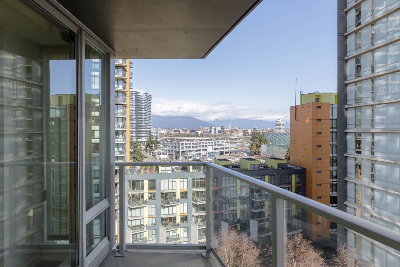View from a high-rise apartment balcony overlooking nearby buildings, with a cityscape and distant mountains under a clear sky.