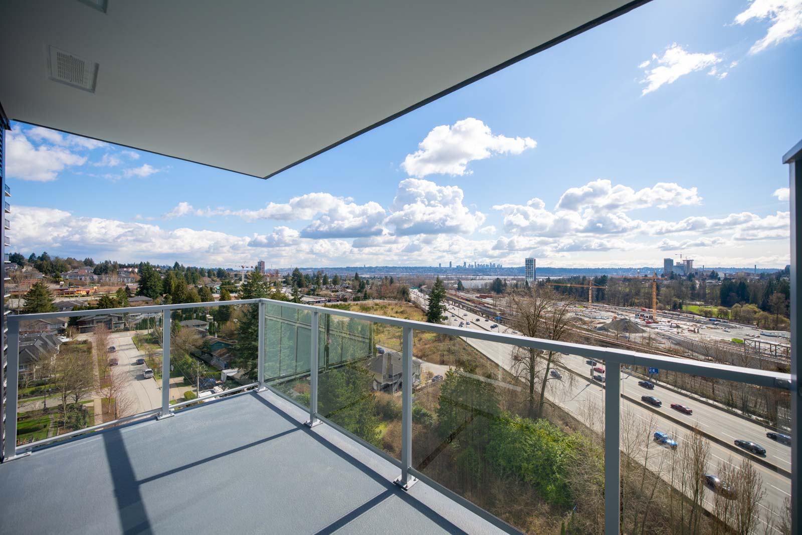 View from a balcony with glass railing overlooking a highway, trees, residential area, and a distant city skyline under a partly cloudy sky.