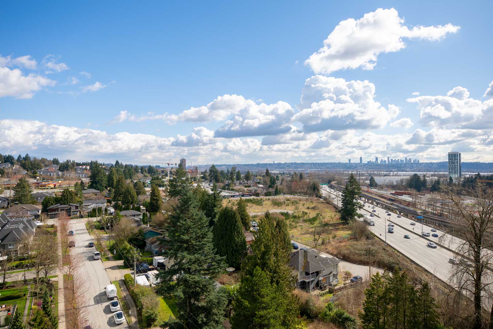 A wide view of a suburban neighborhood with homes, trees, a highway, and a distant city skyline under a partly cloudy sky.