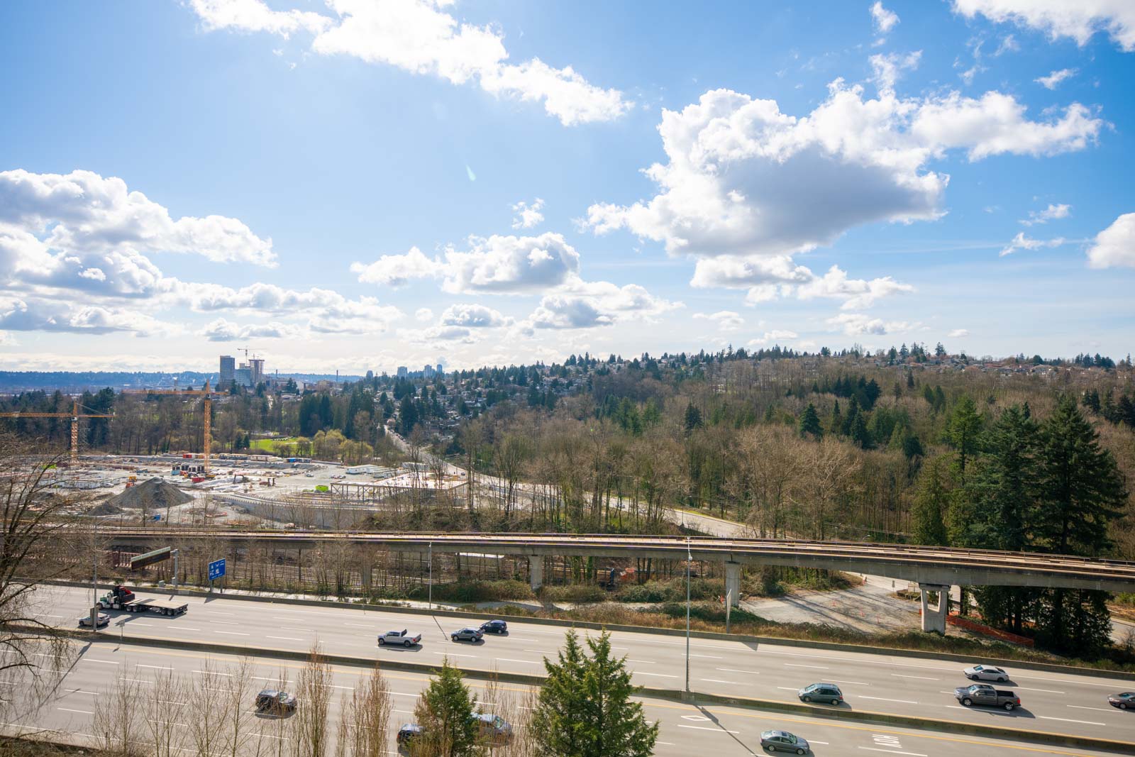 A highway with several cars passes in front of a wooded area under a partly cloudy sky, with buildings visible in the distant background.