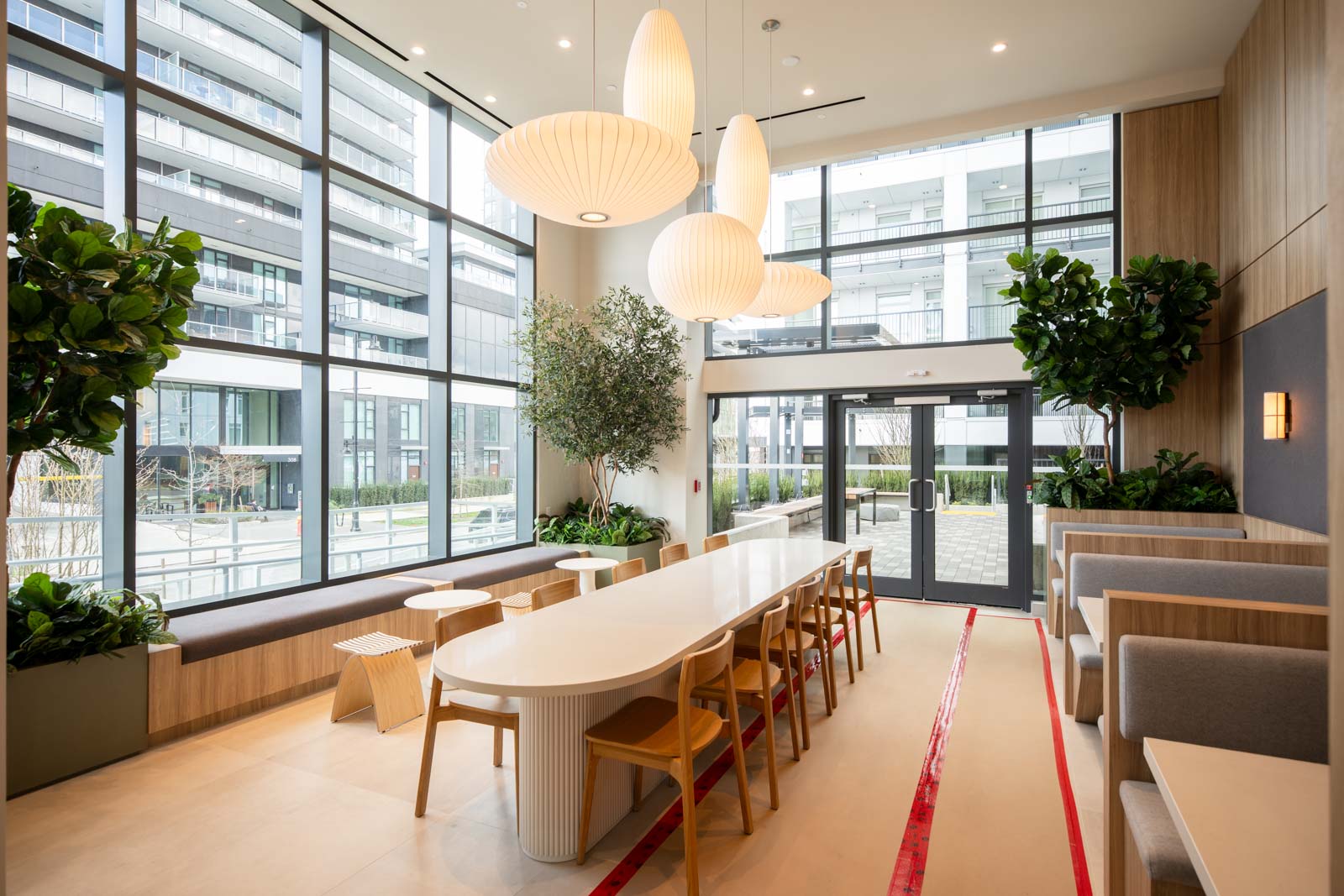 Modern communal dining area with large windows, pendant lights, long table with chairs, booth seating, potted plants, and outdoor view.