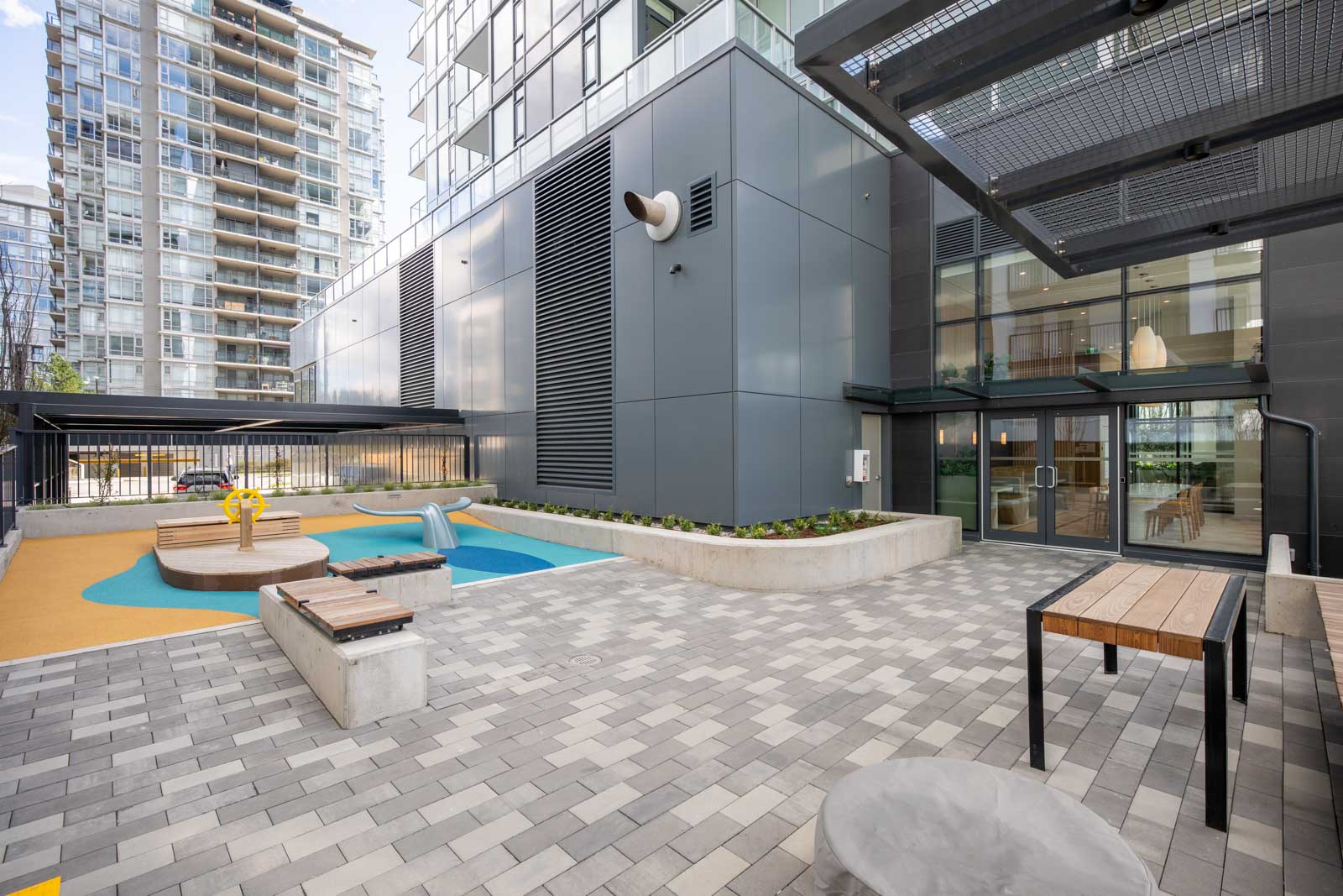 Modern apartment courtyard with paved walkway, built-in planters, benches, tables, and a small children’s play area, surrounded by tall glass buildings.