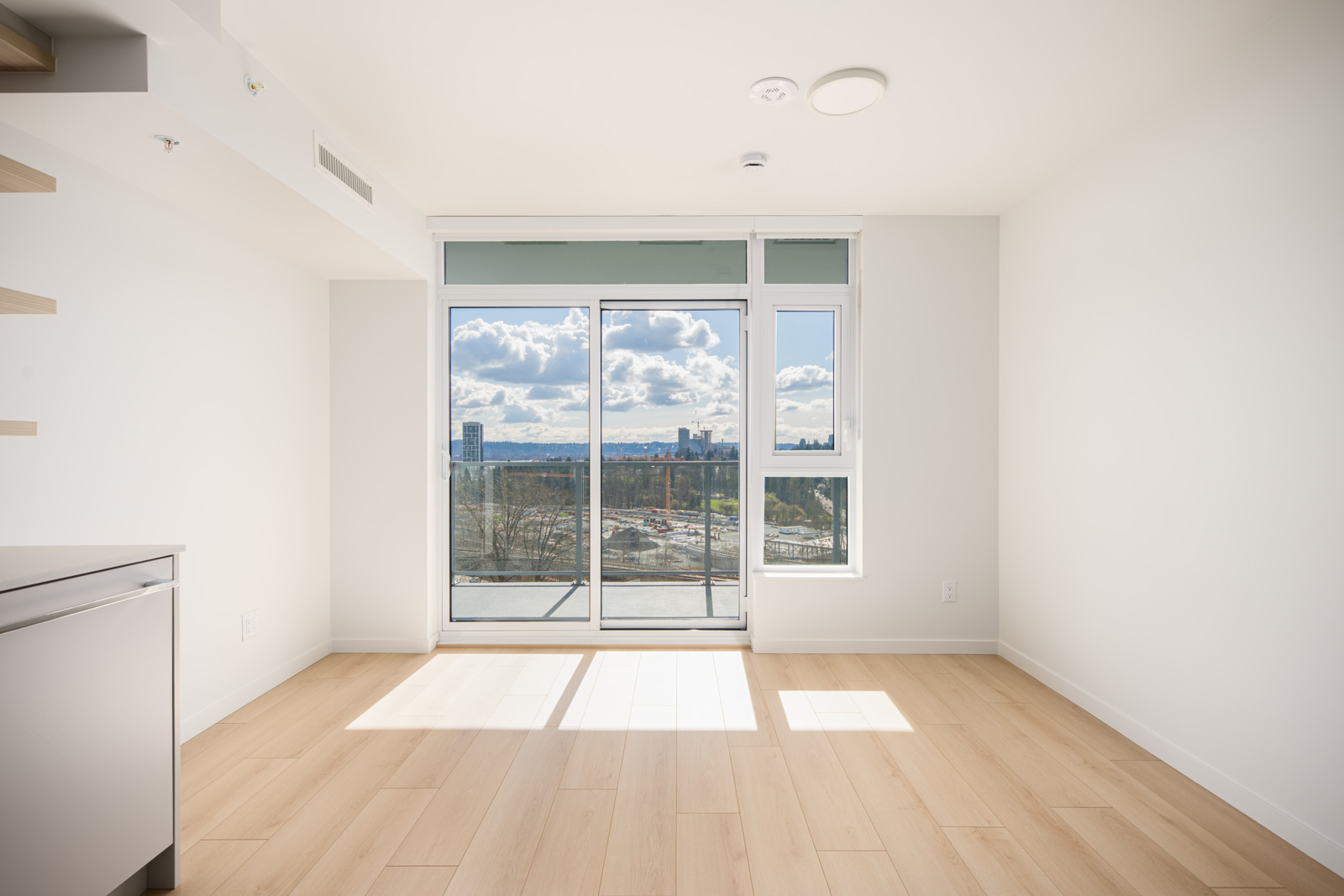 A bright, empty room with light wood flooring, white walls, a large window, and a glass door leading to a balcony with a city view under a partly cloudy sky.