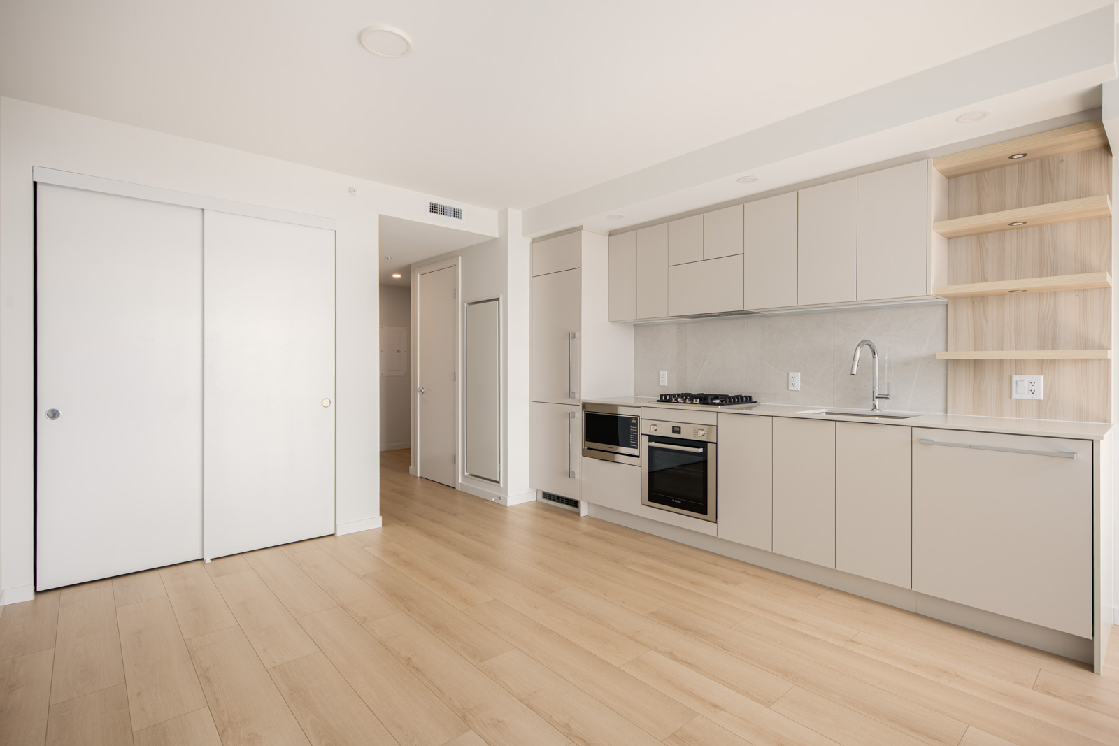 Modern kitchen with light wood flooring, white cabinets, built-in stove and oven, open shelves, and sliding closet doors in a bright, minimal apartment.