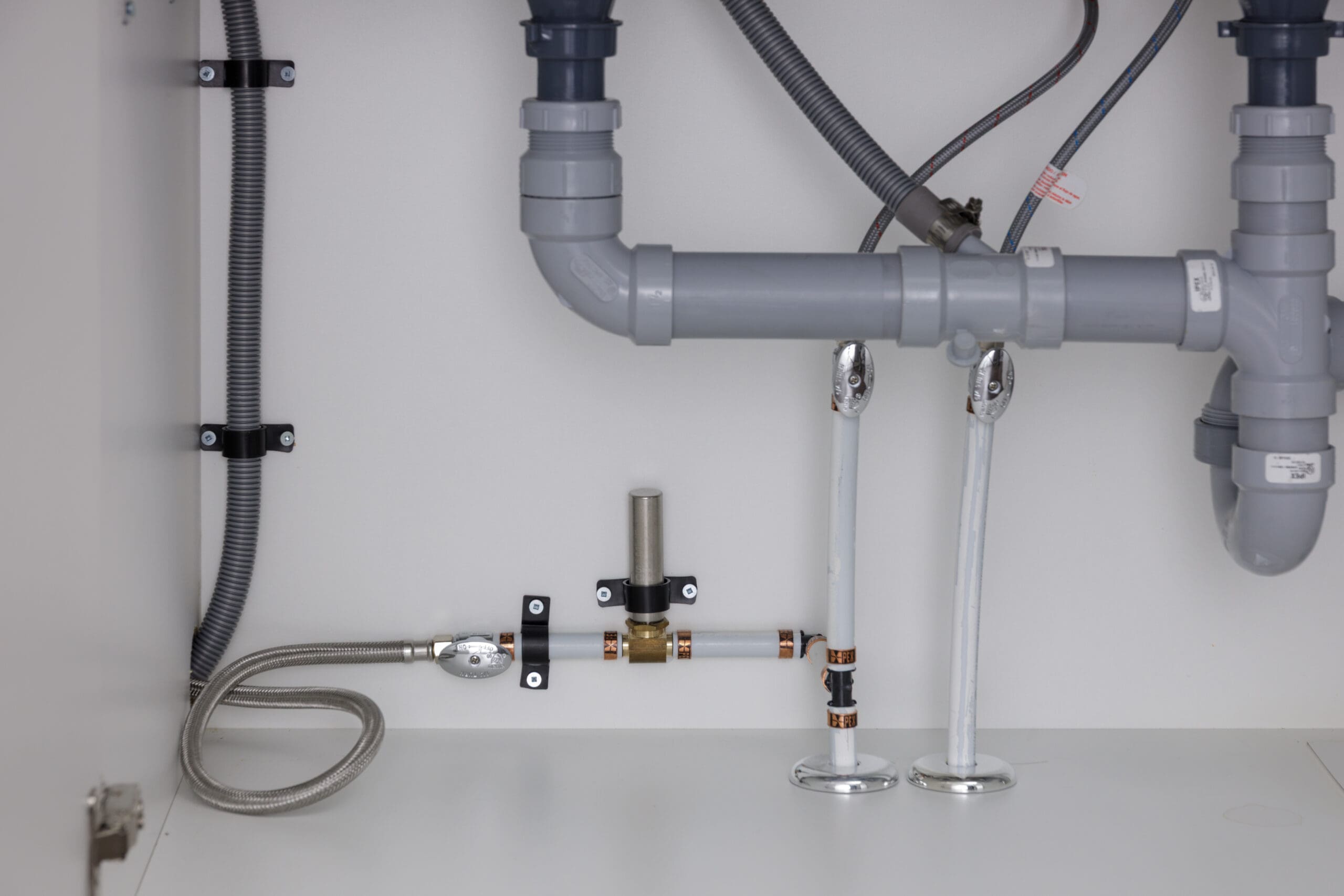 Plumbing pipes, hoses, and fittings under a sink, including water supply lines, drain pipes, and mounting hardware, against a white background.