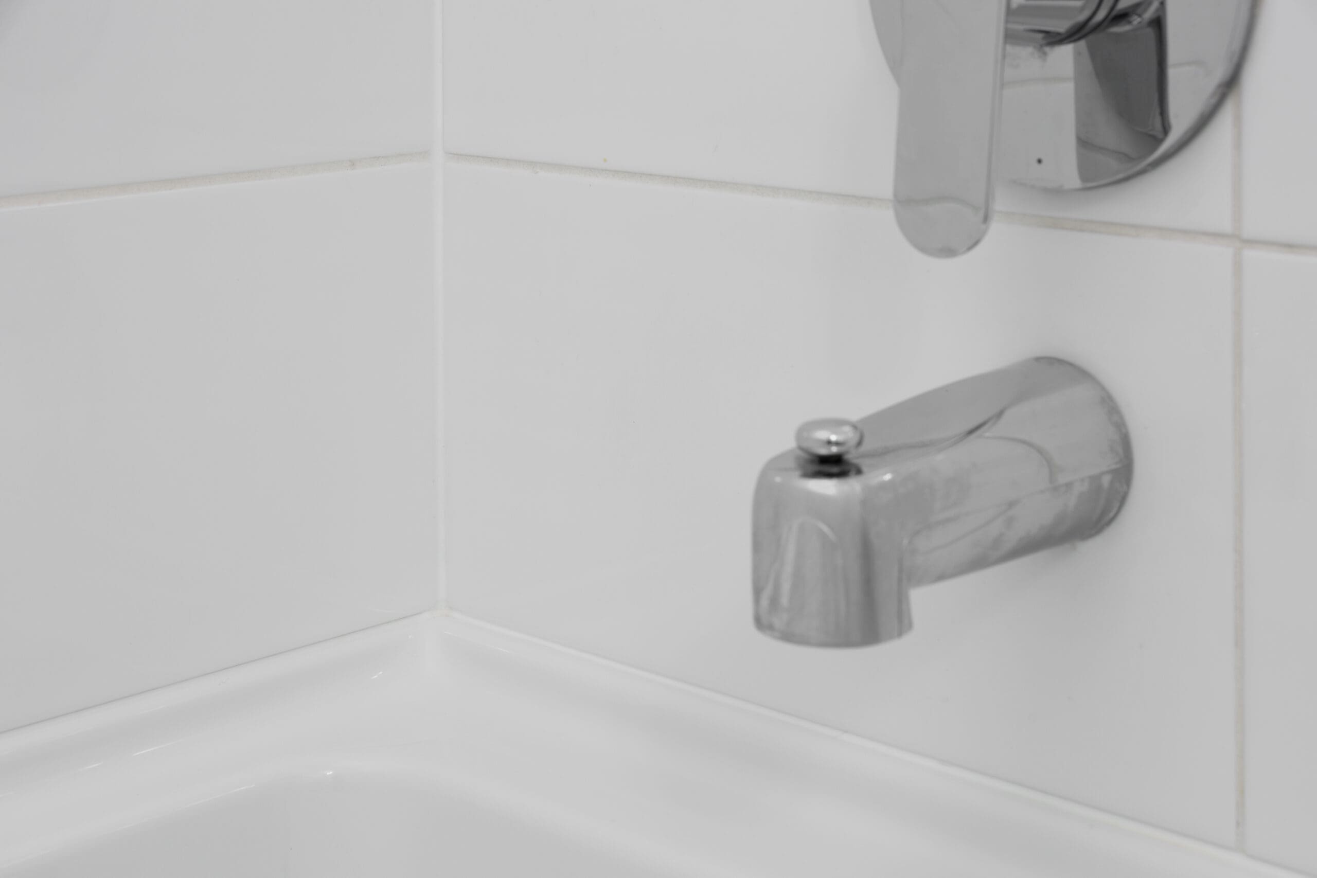 A close-up of a silver bathtub faucet and spout mounted on white tiled bathroom walls above a white bathtub.
