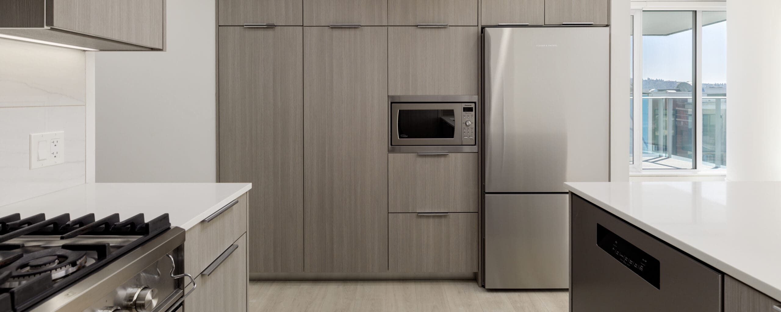 Modern kitchen with light wood cabinets, built-in microwave, stainless steel refrigerator, gas stove, dishwasher, and large window providing natural light.