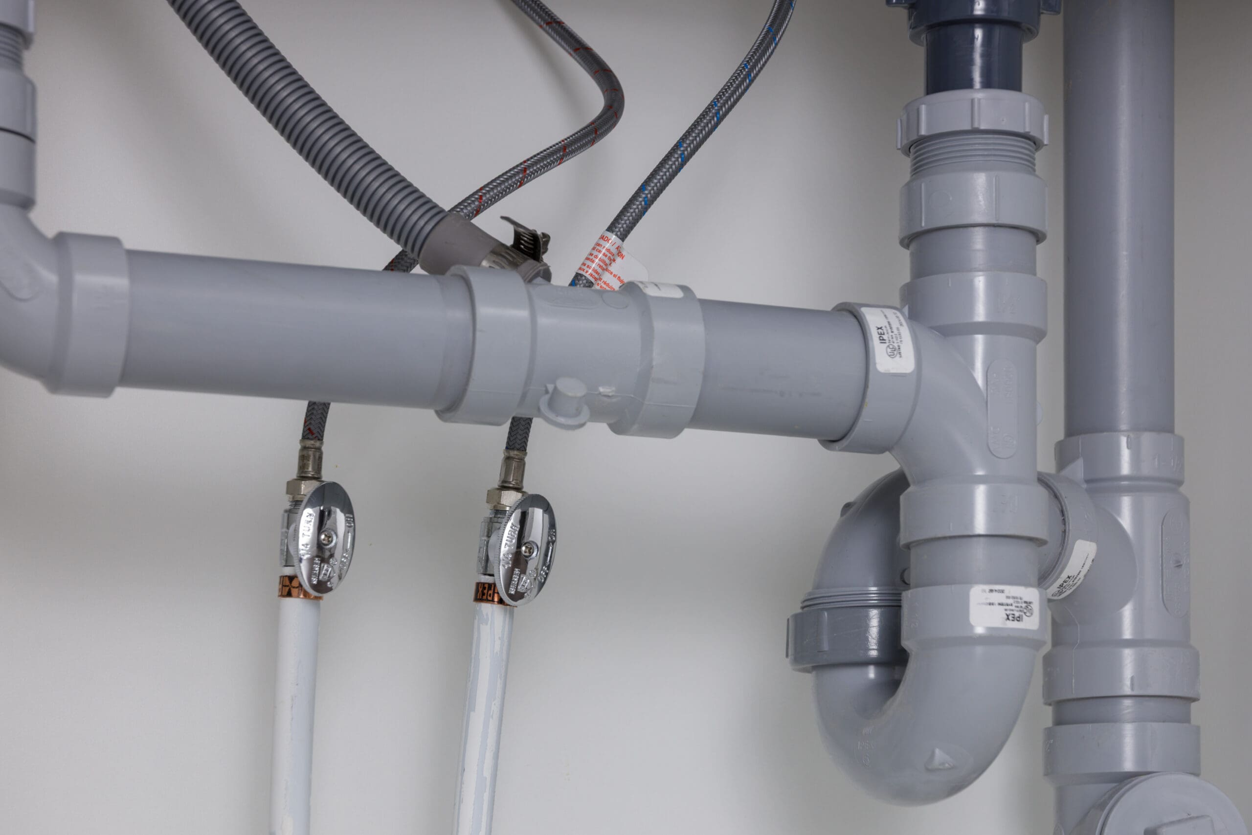 Gray PVC plumbing pipes and fittings under a sink, with two flexible water lines and shut-off valves visible against a white background.