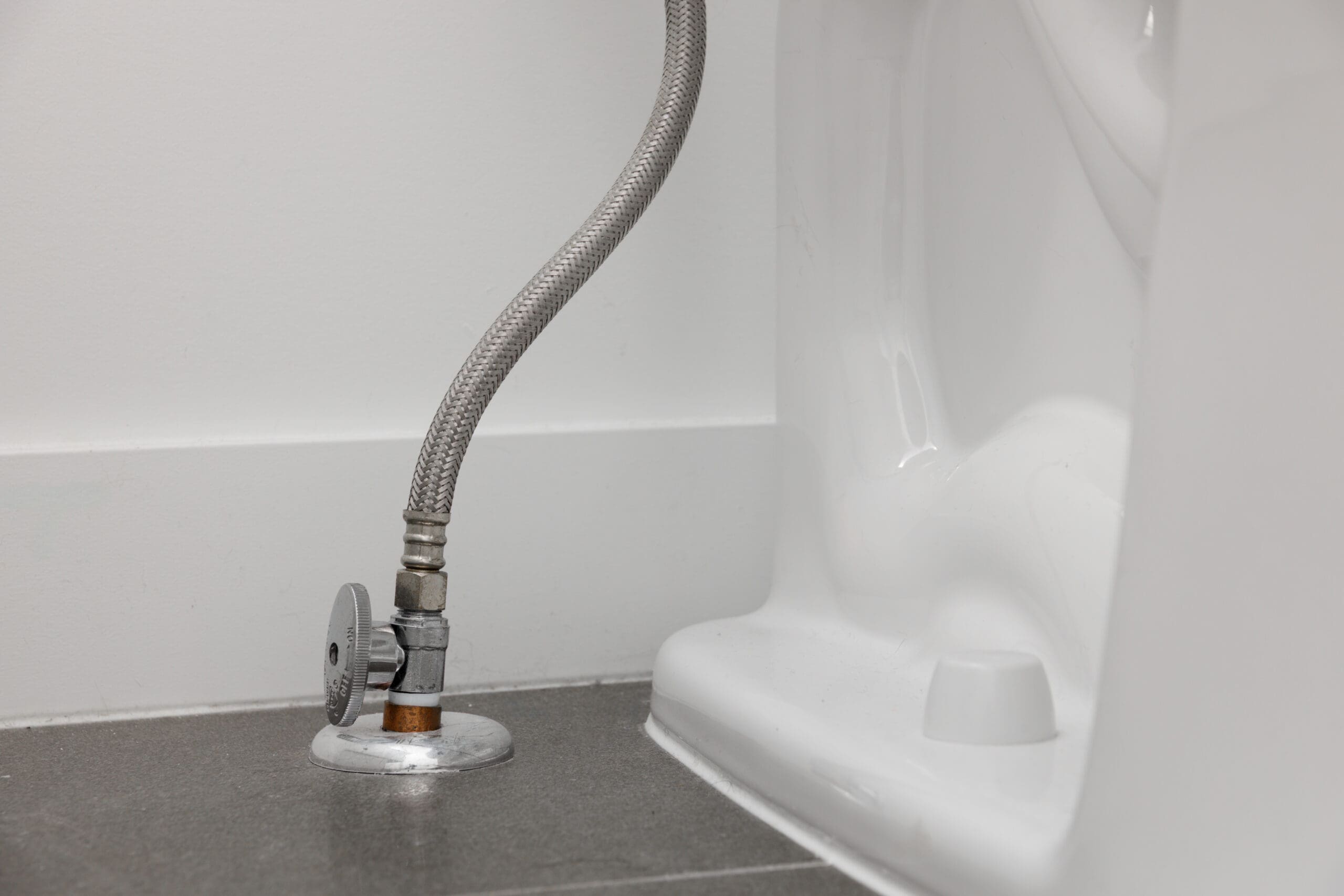 Close-up of a metal water supply line connected to a shut-off valve at the base of a toilet on a tiled bathroom floor.