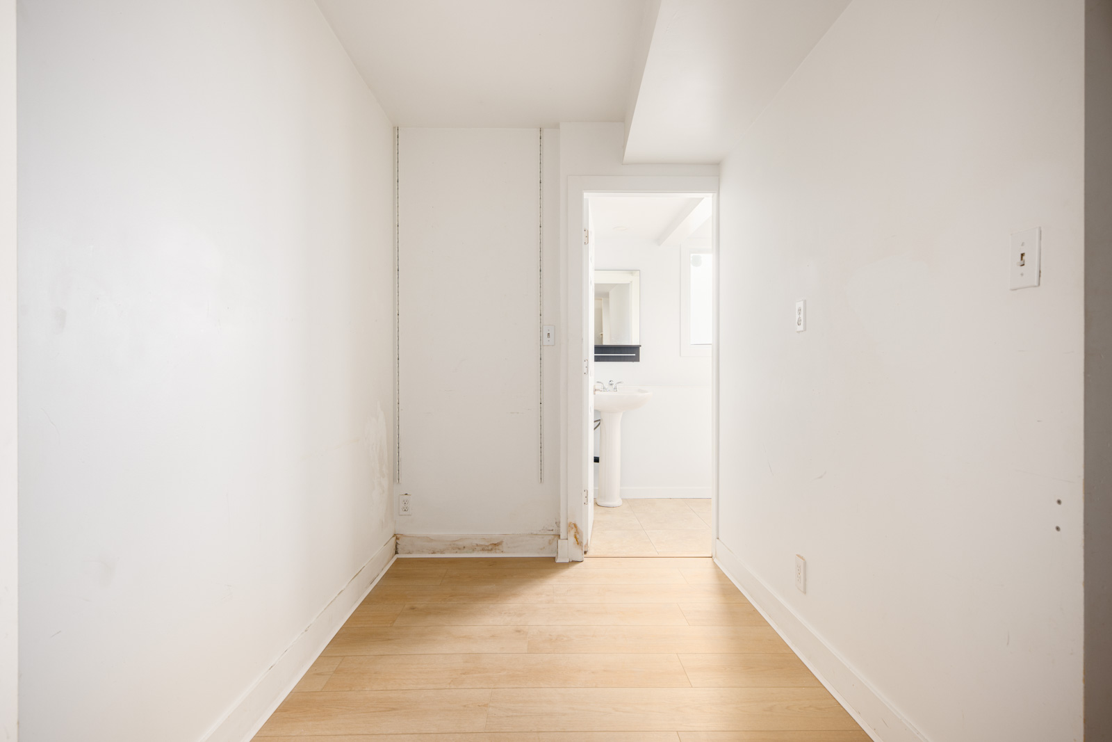 A small, empty room with white walls, light wood flooring, and a door leading to a bathroom with a sink and mirror visible.
