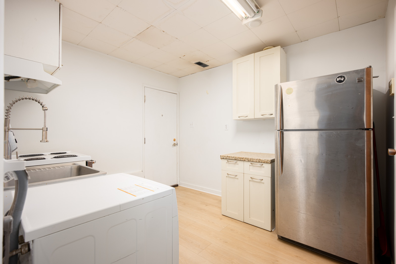 Small kitchen with light wood floor, white walls, washing machine, stove, stainless steel refrigerator, and white cabinets under a stained ceiling.