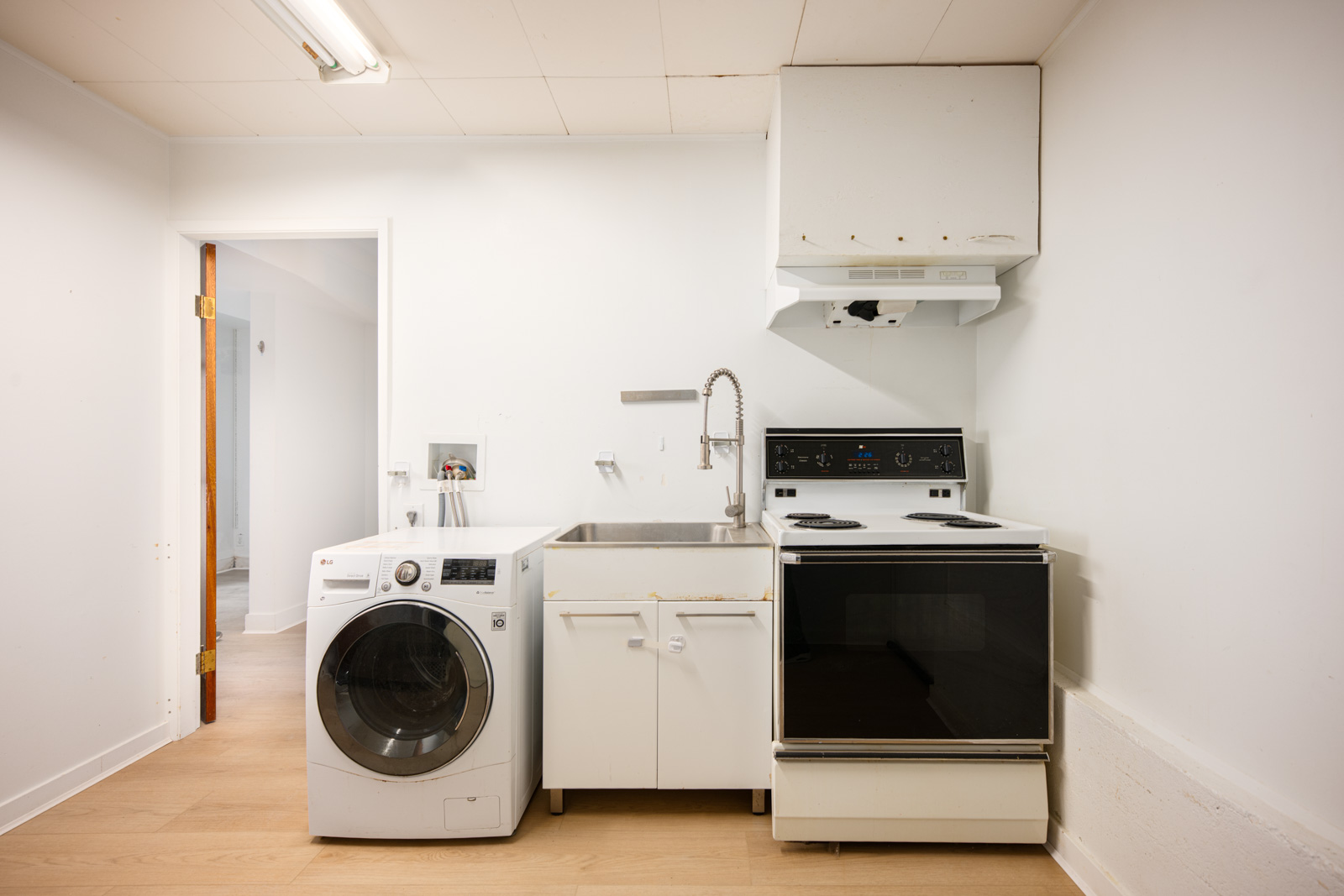 A small utility room with a washing machine, utility sink, and an electric stove with a range hood, all set against white walls and a light wood floor.
