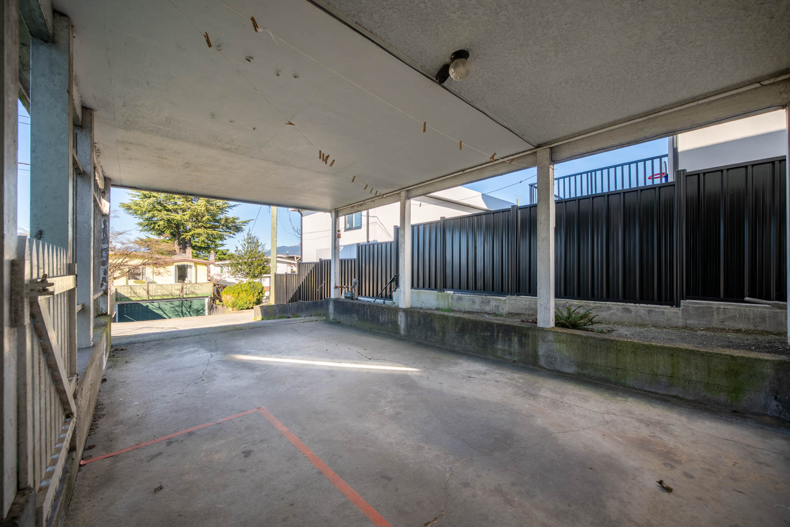 Covered concrete patio area with a few clotheslines and clothespins overhead, surrounded by black metal fencing and neighboring buildings visible in the background.