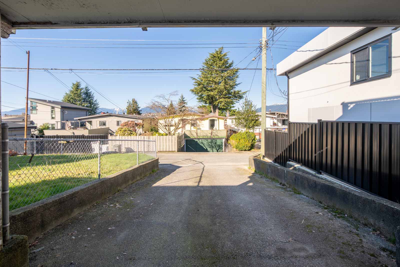 View from a driveway looking out toward a residential street, with houses, a fenced yard, trees, and mountains in the background under a clear blue sky.