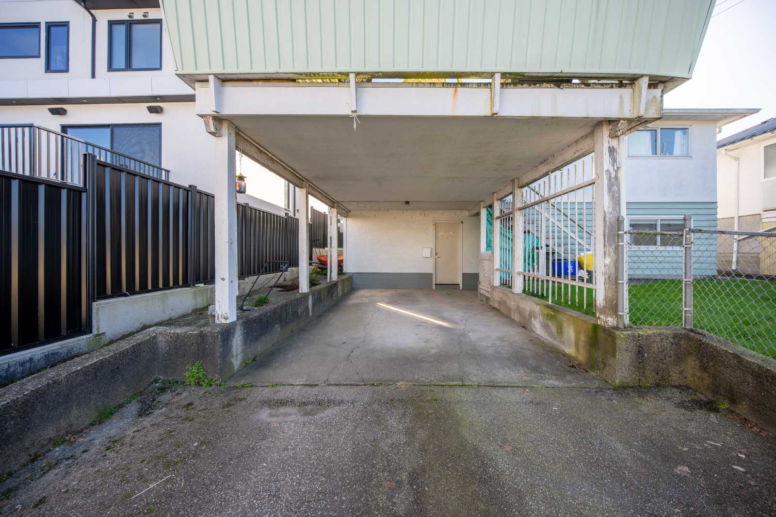 A covered carport with concrete flooring, surrounded by a black metal fence on one side and a chain-link fence on the other, next to residential buildings.