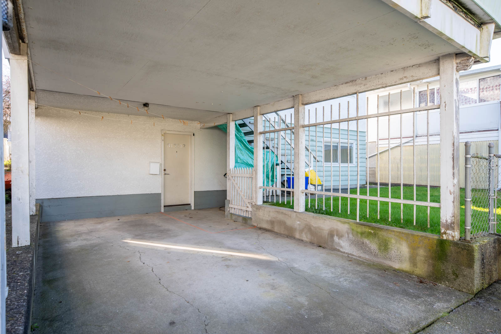 Covered outdoor parking area with concrete floor, adjacent to a gated yard and staircase, next to a door leading into a building.
