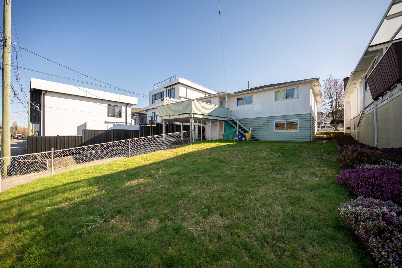 A two-story house with a sloped green lawn, chain-link fence, and neighboring modern houses under a clear blue sky.