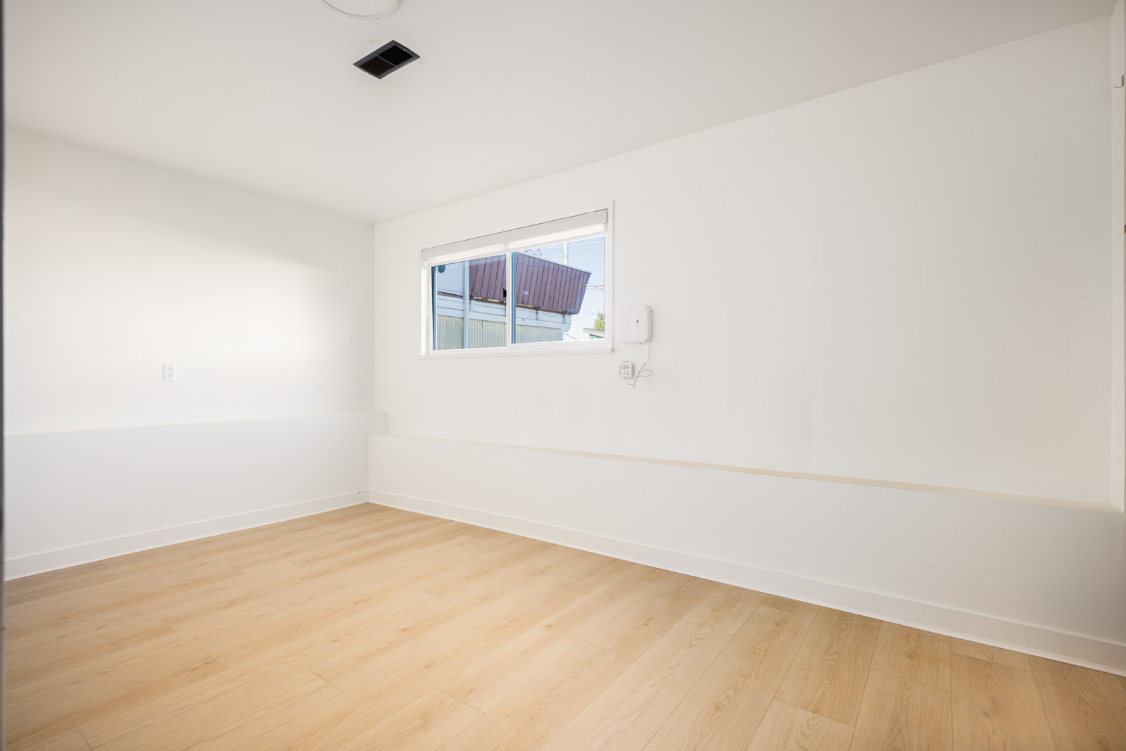 Empty room with white walls, light wood flooring, a single window, and a wall-mounted device below the window.