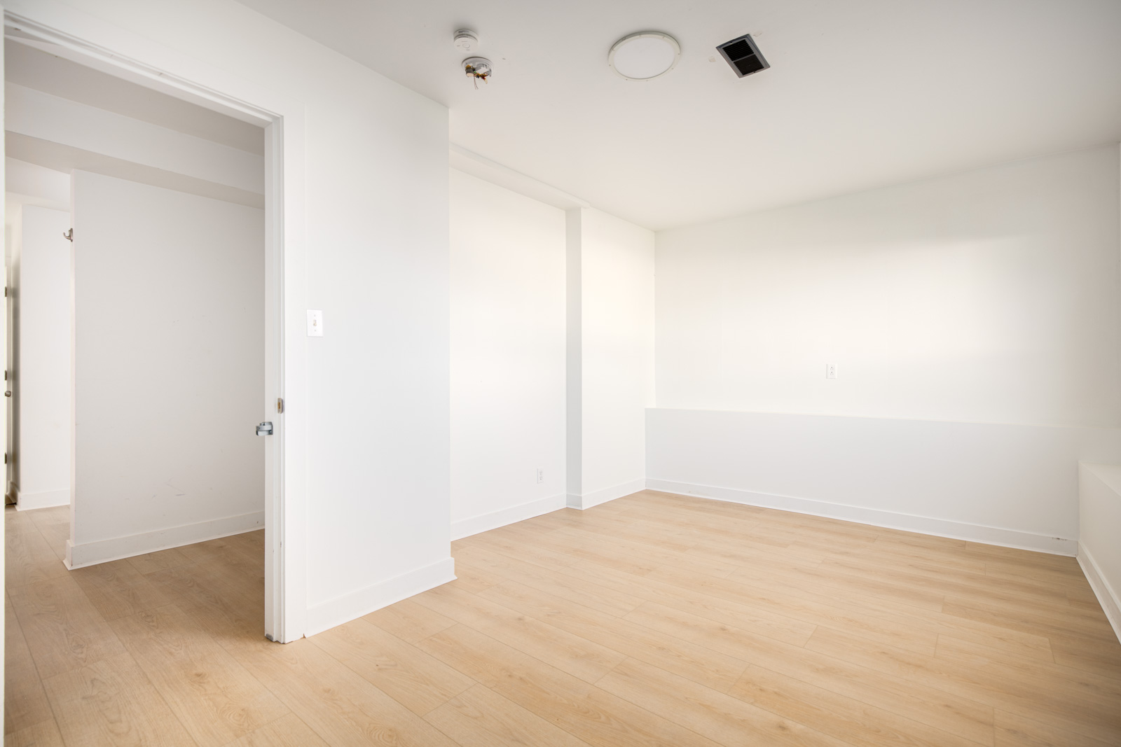 Empty room with white walls, light wood flooring, an open door to a hallway, ceiling fixtures, and a wall vent.