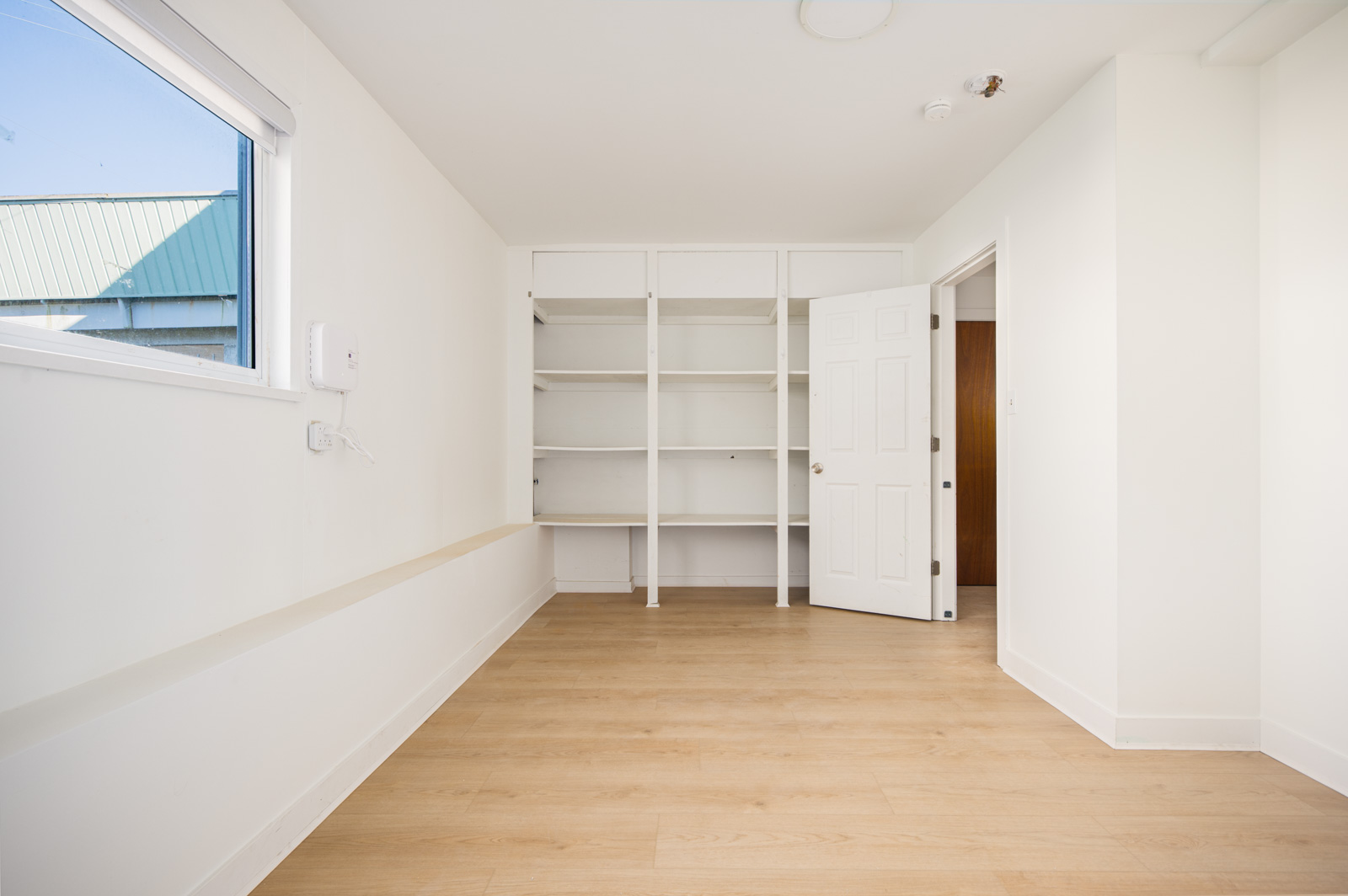 Empty room with light wood flooring, white walls, open closet shelves, an open door, and a window letting in natural light.