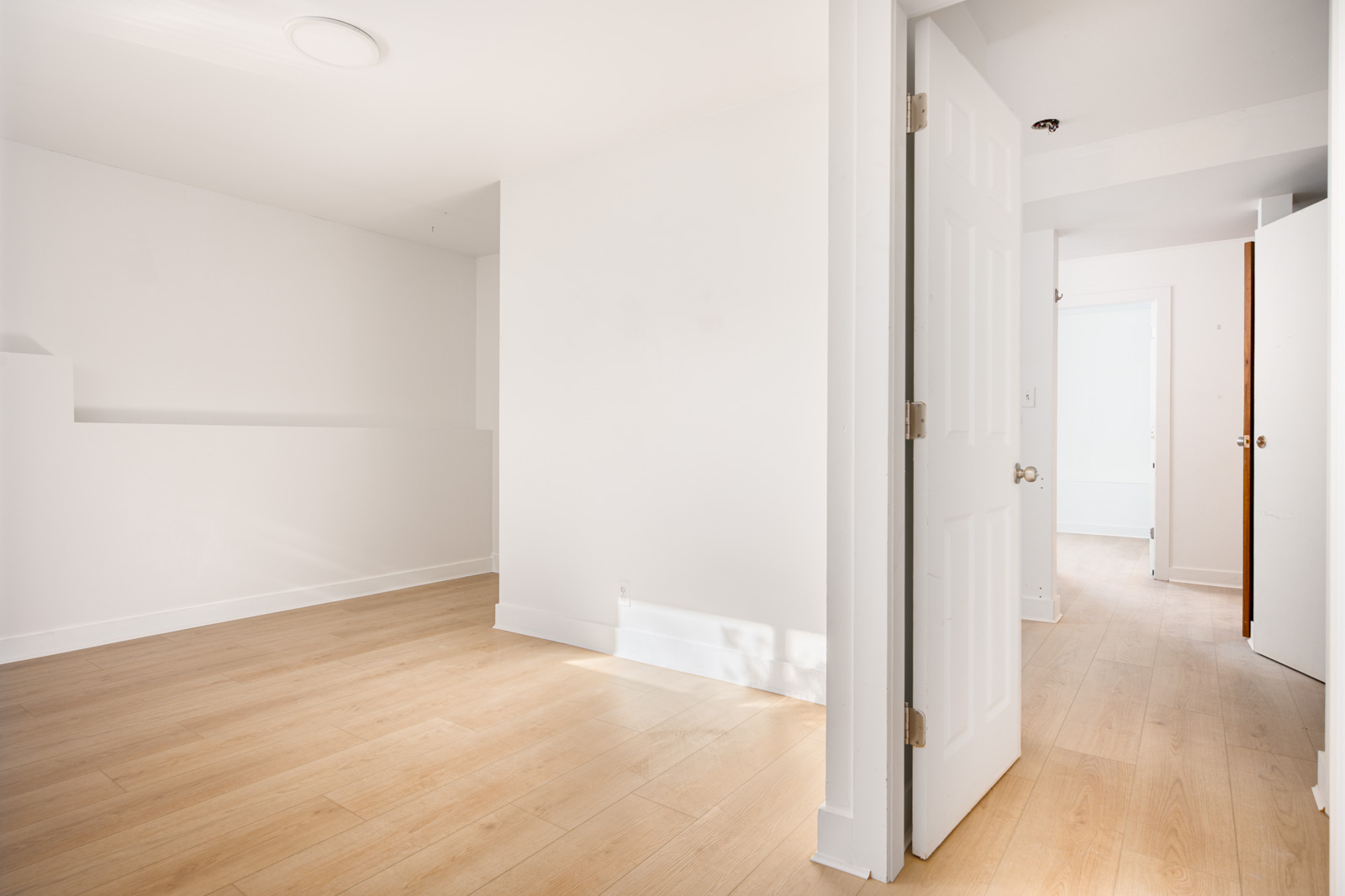Bright, empty room with light wood flooring, white walls, and several open white doors, allowing natural light to enter.