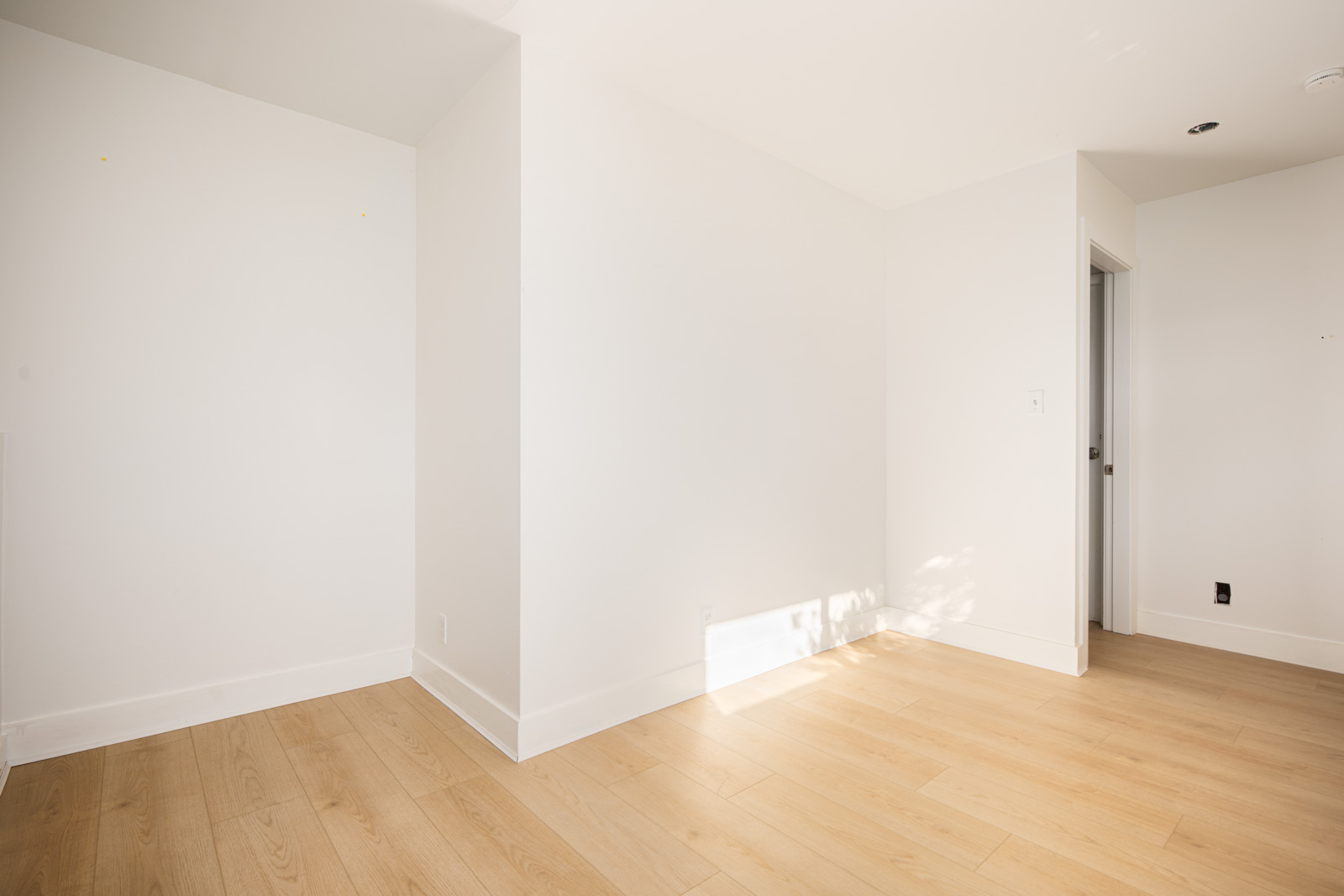 Empty room with light wood flooring, white walls, and an open doorway leading to another area; sunlight casts shadows near the corner.