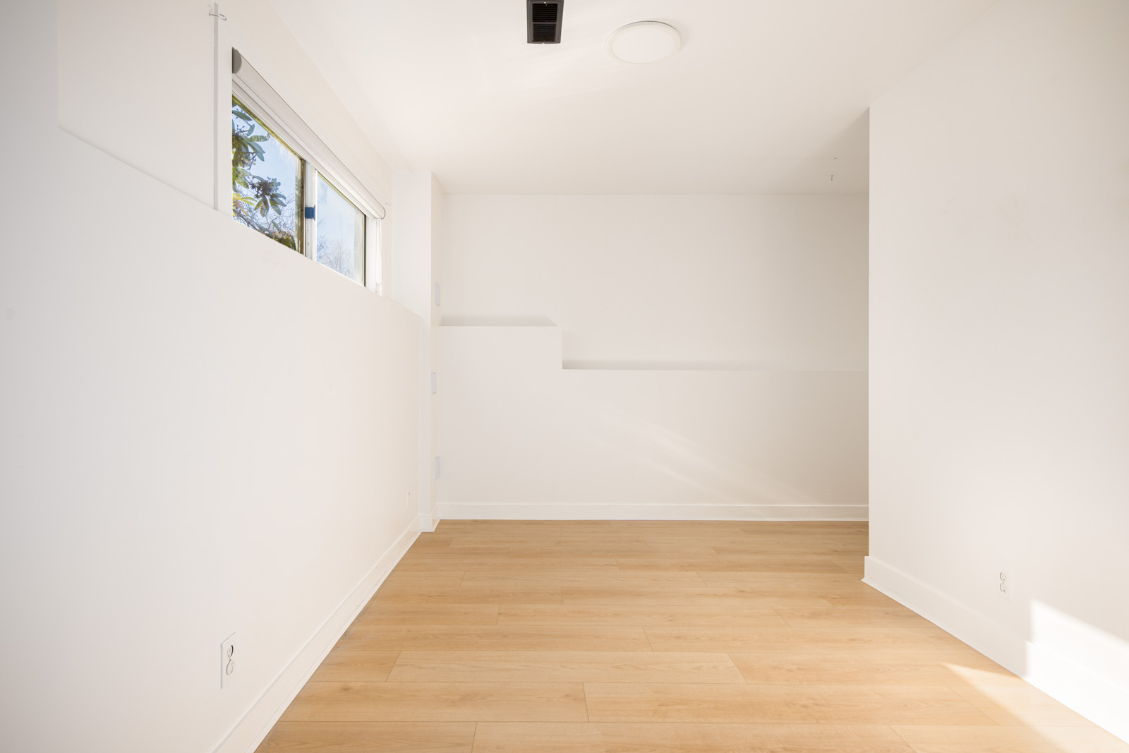 Empty, unfurnished white room with light wood flooring, a large window on the left wall, and a recessed shelf built into the back wall.