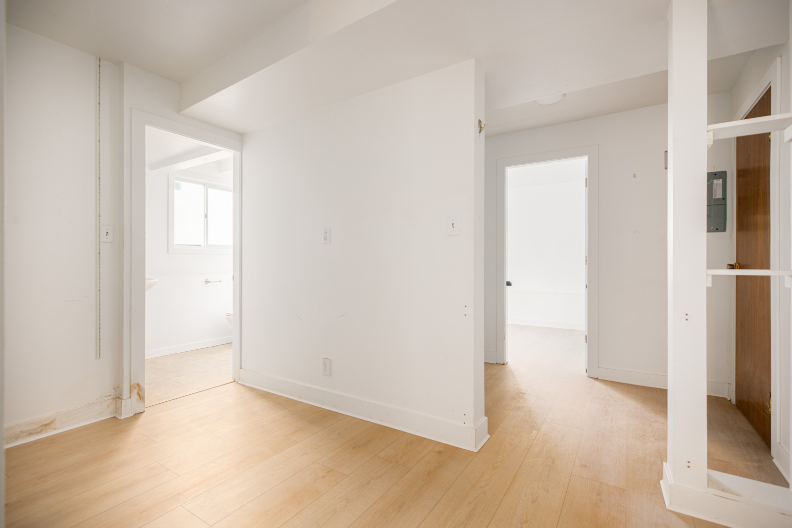 A bright, empty room with light wood flooring, white walls, and three doorways leading to other rooms, including a visible bathroom.