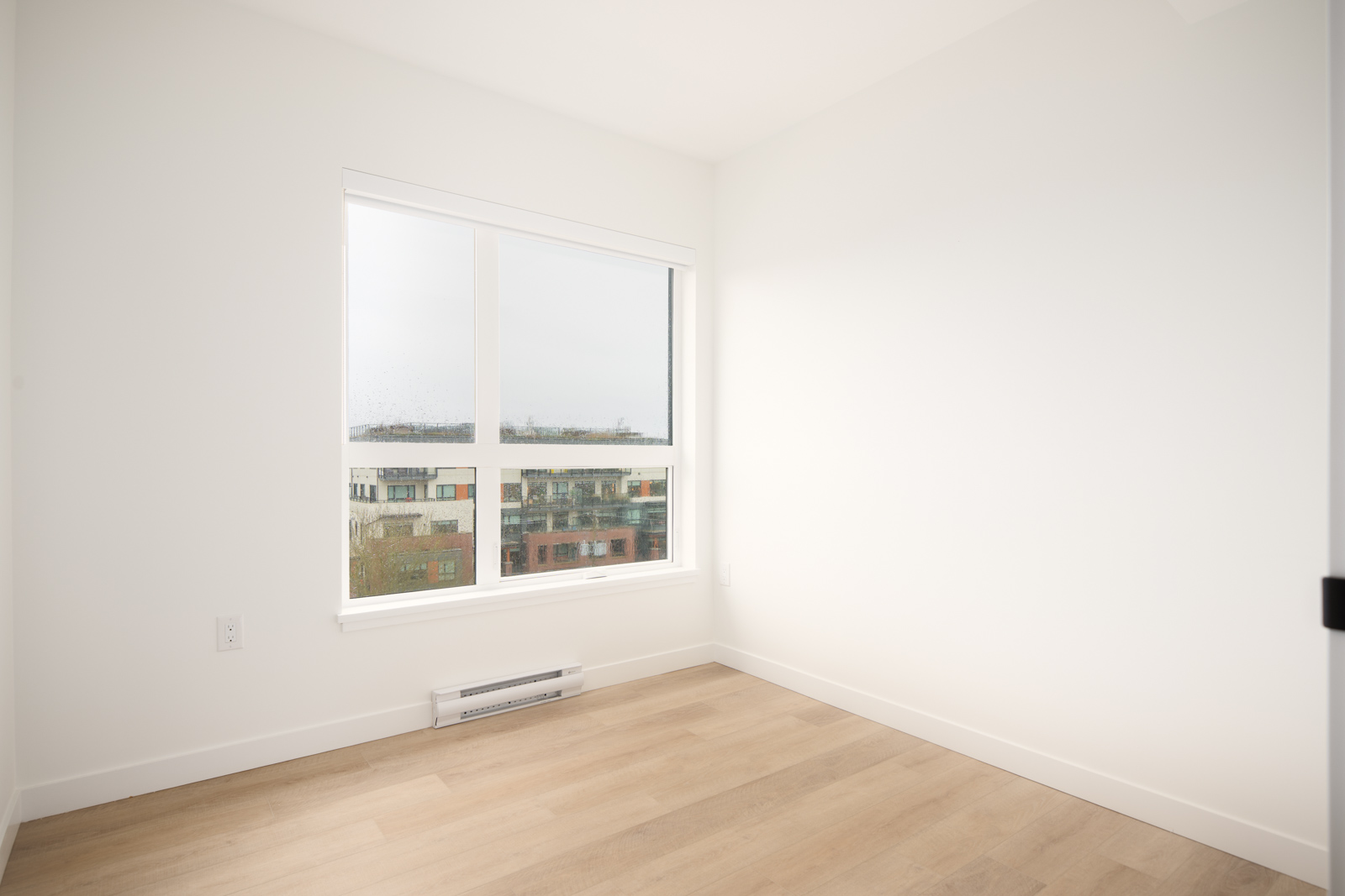 Empty, unfurnished room with white walls, light wood flooring, a large window, and a view of apartment buildings outside.