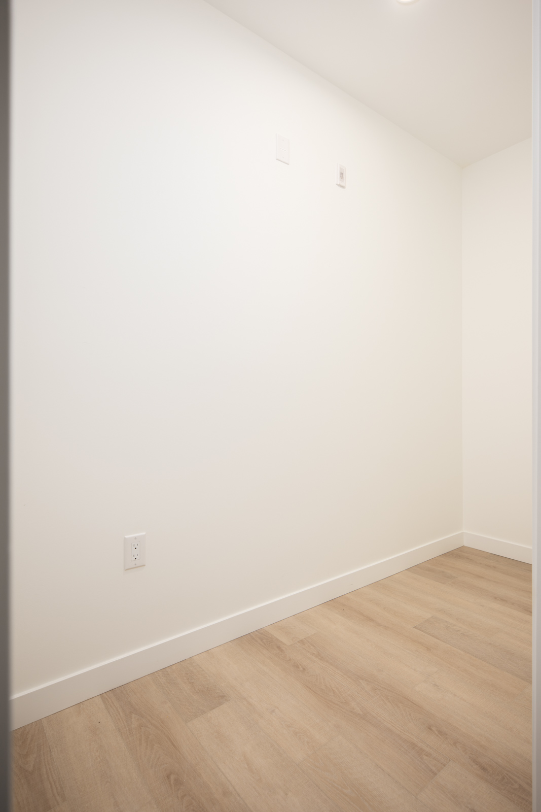 Empty room with white walls, light wood flooring, a power outlet, and two small wall fixtures near the ceiling.