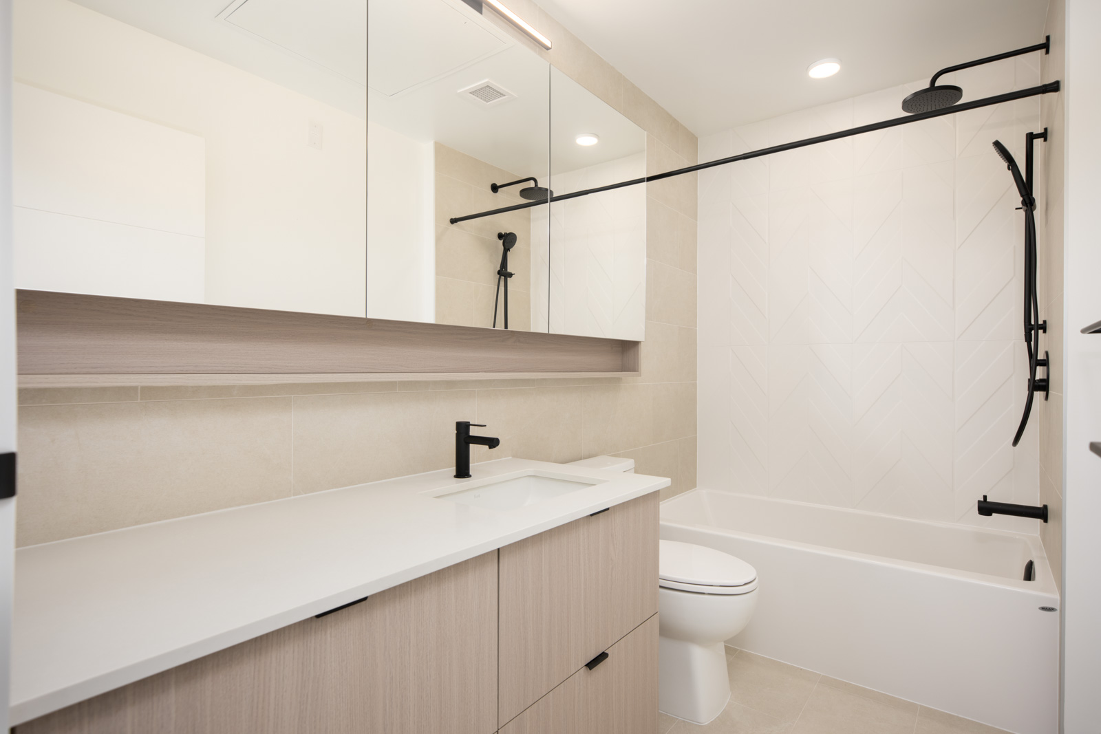 Modern bathroom with a light wood vanity, white countertop, black fixtures, large mirror, and a bathtub with a black showerhead and white tile wall.