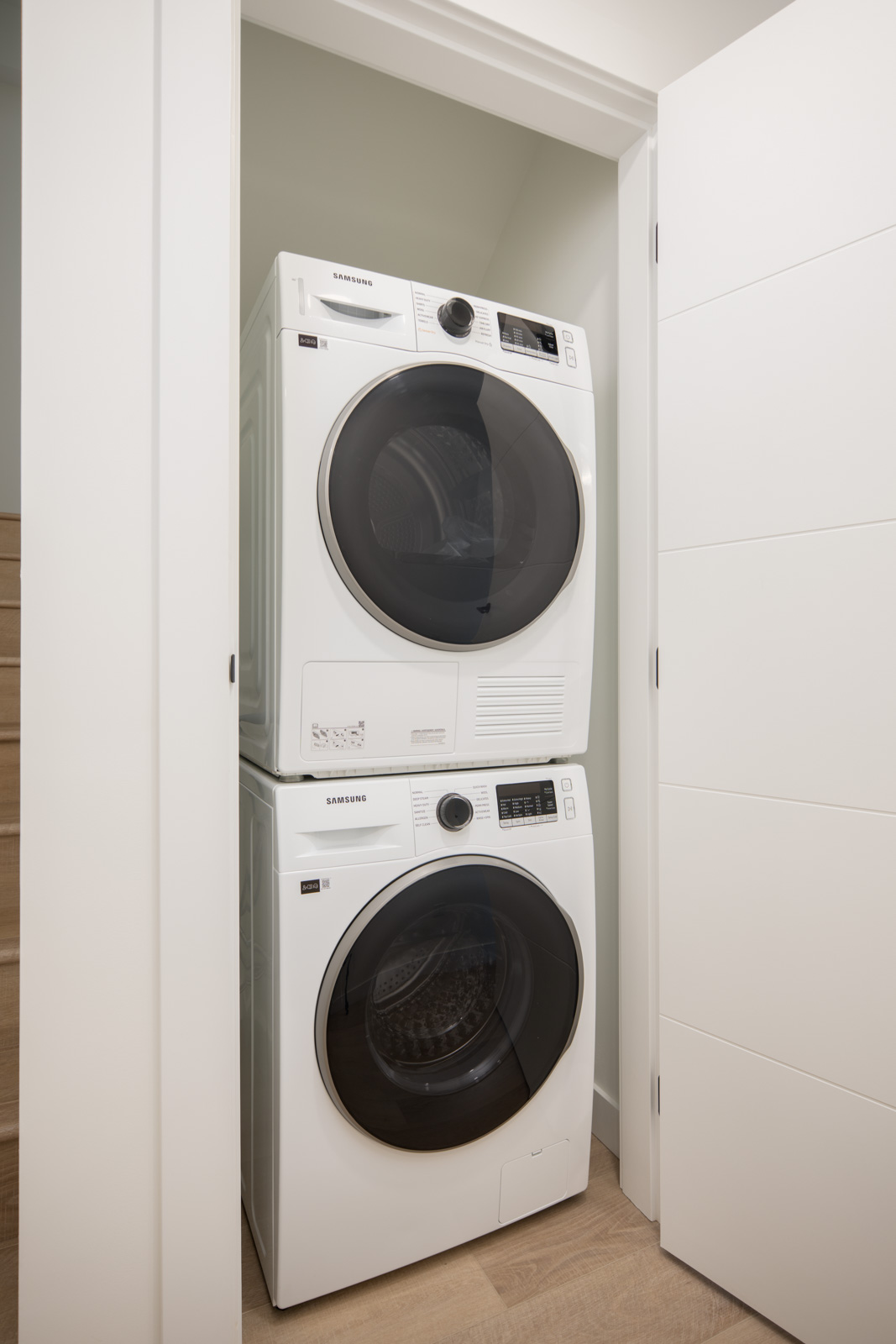 Stacked white Samsung washer and dryer units are placed in a closet with white double doors, next to a wooden staircase.