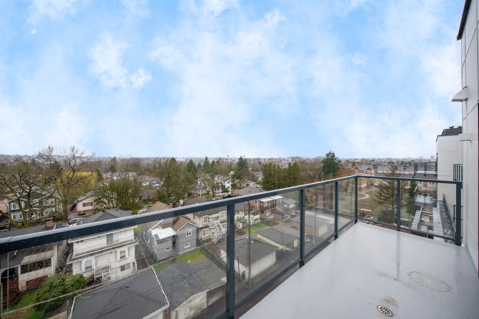 View from a high balcony with a glass railing overlooking residential houses, trees, and a cloudy blue sky in the distance.