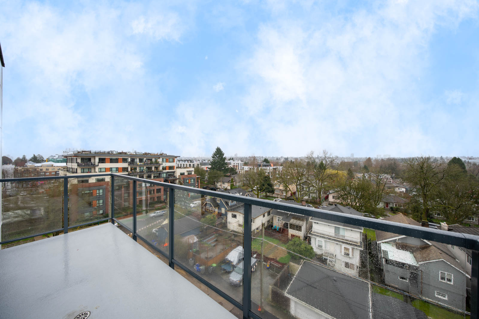 View from a balcony with glass railing overlooking residential houses, trees, and mid-rise apartment buildings under a partly cloudy sky.