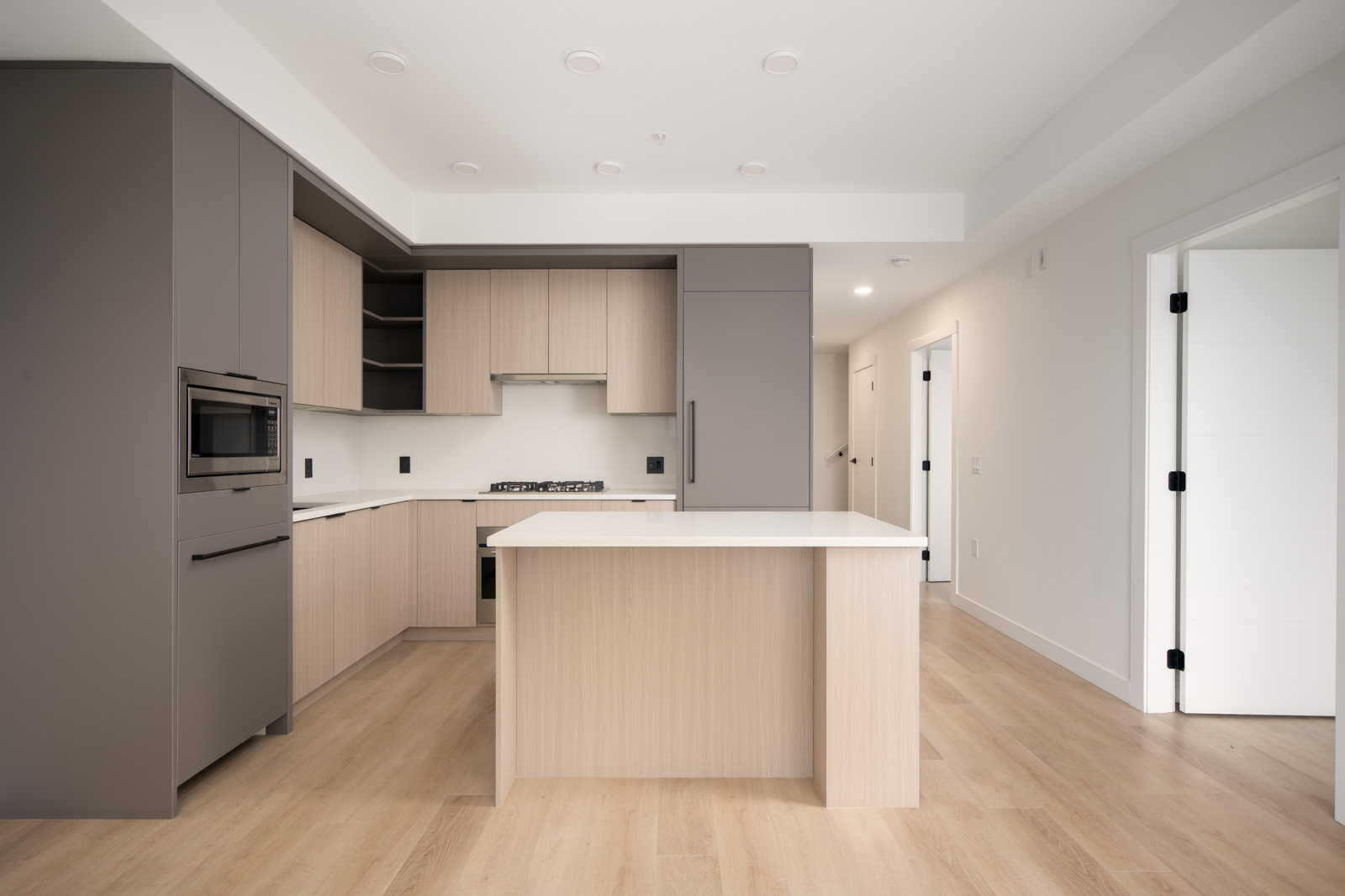 Modern kitchen with light wood floors, beige and gray cabinets, built-in oven, gas stove, center island, and white walls. Doorways visible in the background.