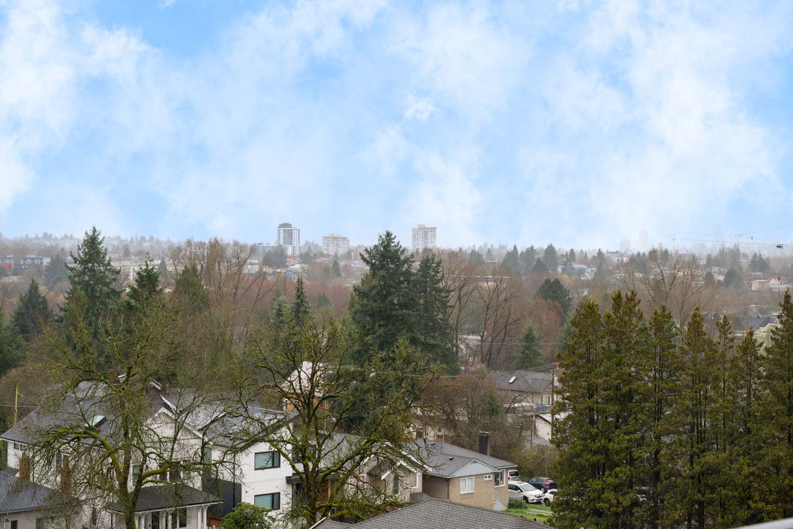 Suburban neighborhood with houses, trees, and a distant view of mid-rise buildings under a partly cloudy blue sky.