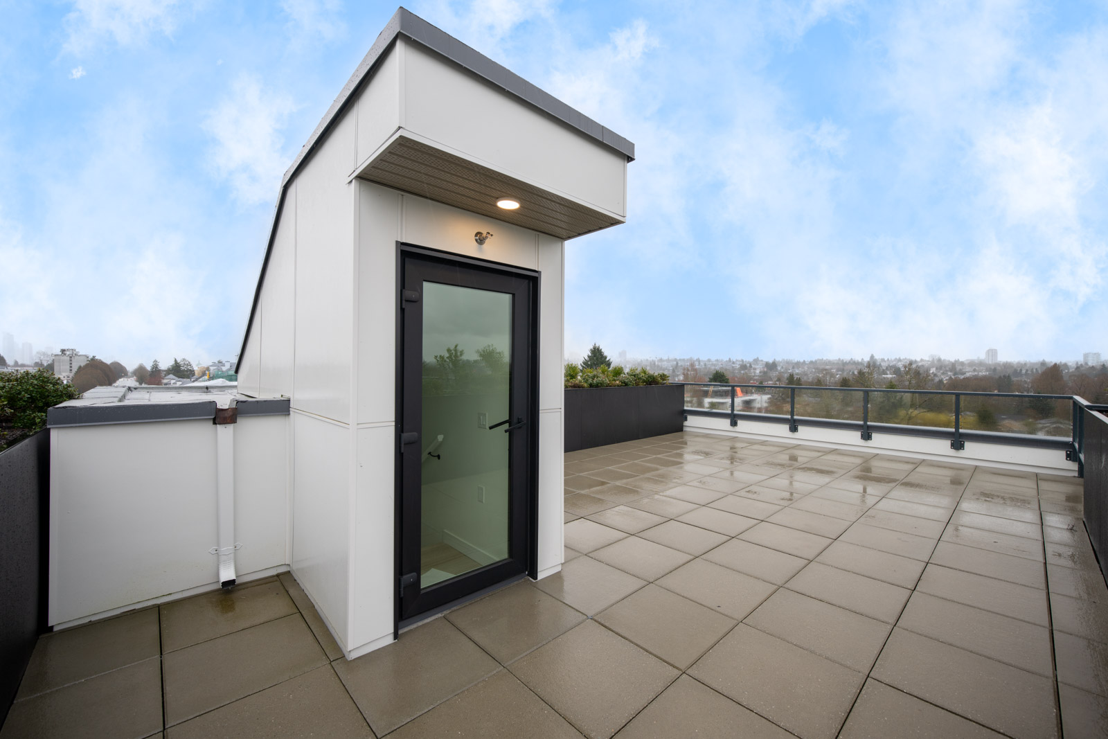 Rooftop terrace with tiled flooring, black metal railing, and a small white entry structure with a glass door, under a partly cloudy sky.